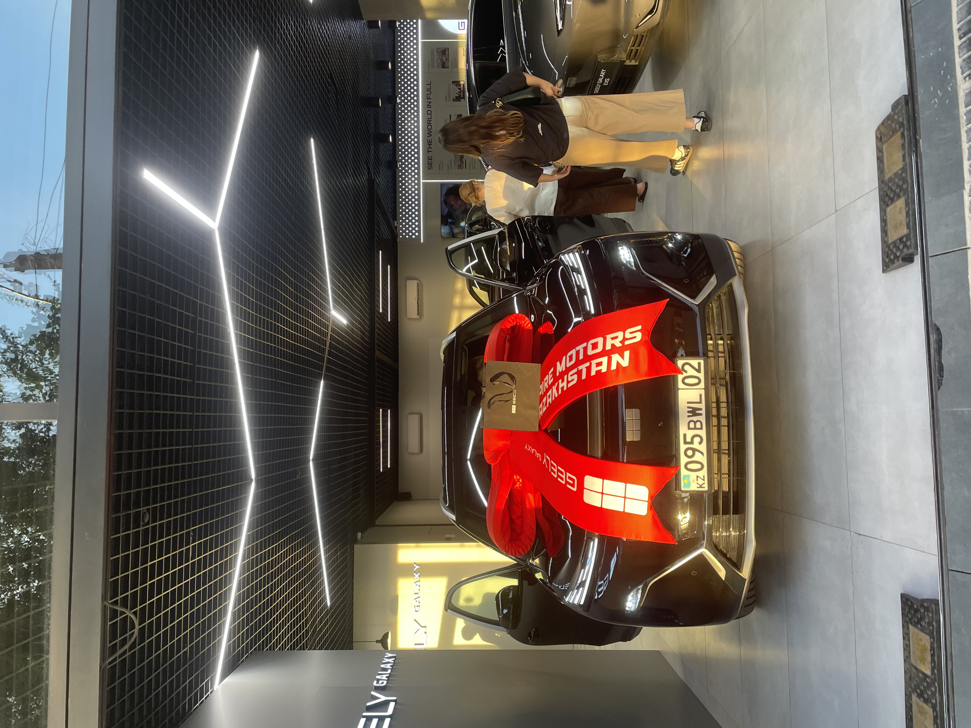 In a modern car dealership in Almaty, Kazakhstan, a new, dark-colored Geely Galaxy SUV is presented with a large, red bow on its hood. The car's doors are open, and its headlights and license plate are illuminated. The license plate reads "KZ 095BWL 02." Two women, one wearing a white shirt and brown pants, and another in a dark t-shirt and beige pants, stand near the car, seemingly examining it. A third person's silhouette is visible through the open driver's side door. The dealership's interior features a grid ceiling with integrated LED lighting and a "GEELY GALAXY" logo prominently displayed on the wall. The floor is tiled in a light gray color. Another car, possibly a Geely Galaxy, is partially visible to the right of the featured vehicle. The overall impression is one of a new car handover or display.