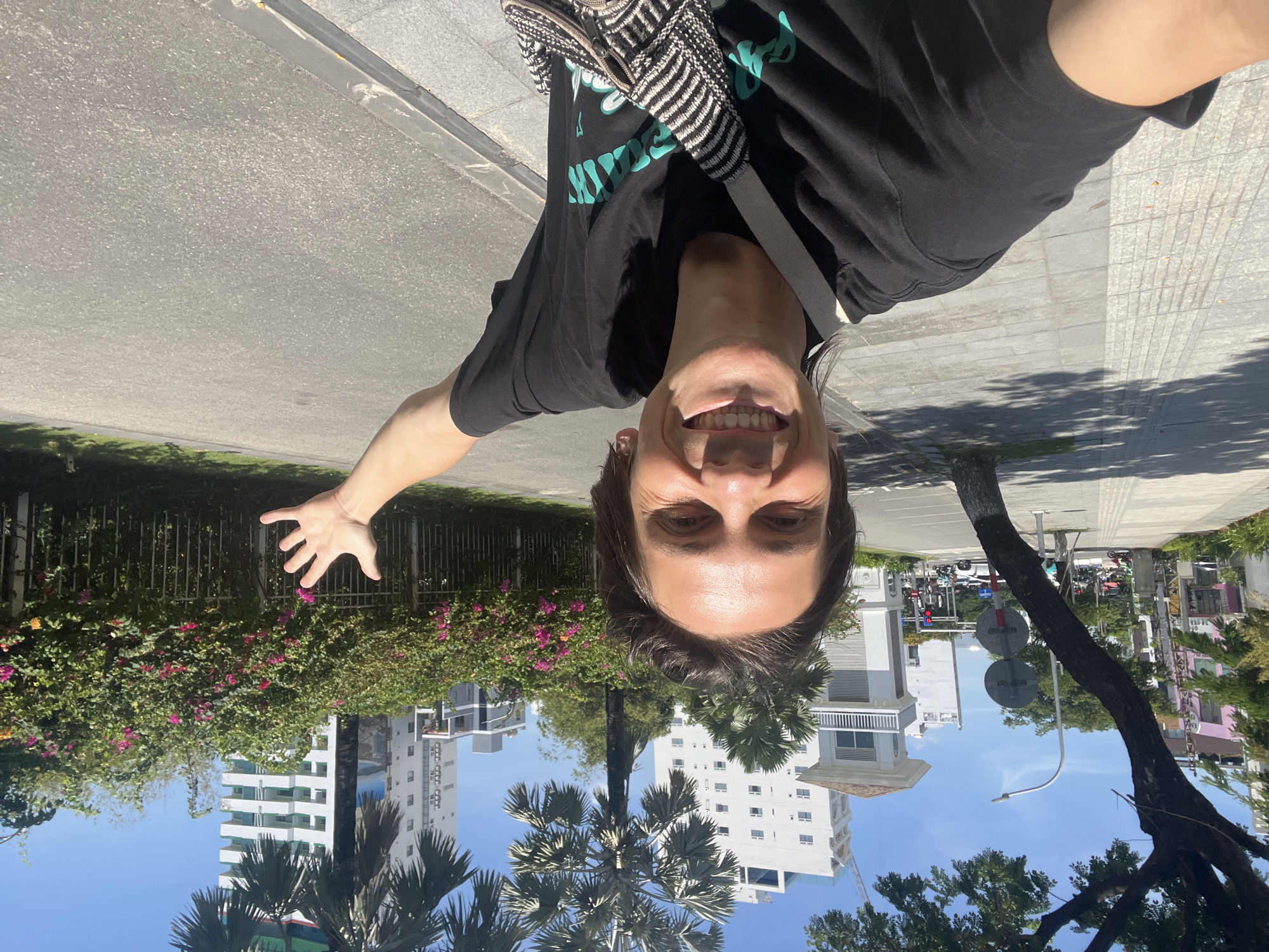 A man is taking a selfie outdoors on a sunny day. He is wearing a black t-shirt with a graphic design and a crossbody bag with a black and white striped pattern. His right arm is extended to the side, with his hand open. He is smiling broadly at the camera.
The background shows a city street with a sidewalk. There are trees, including a palm tree, and buildings in the distance. The buildings appear to be modern, with white exteriors and balconies. There is also a section of lush green foliage with pink and orange flowers along a fence. The street is paved with asphalt, and the sidewalk is made of light-colored paving stones. The man appears to be in Đà Nẵng, Vietnam, given the architecture and vegetation.