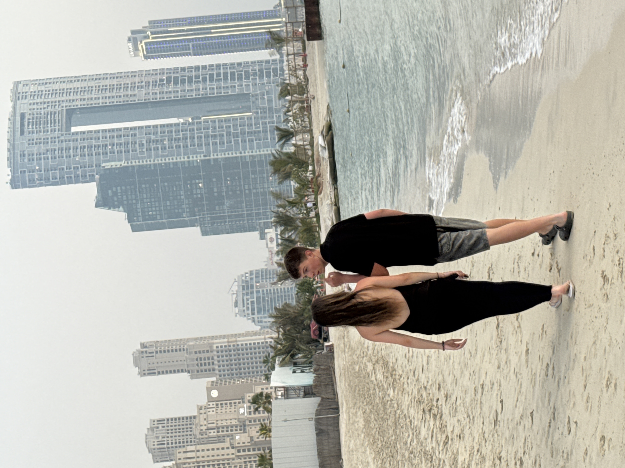 The image captures a beach scene with a couple in the foreground and a cityscape in the background. The couple, a young man and woman, are standing on the sand near the water. The man is wearing a black t-shirt and grey shorts, and the woman is in a black dress. They appear to be interacting, with the man looking at the woman. The water is calm with gentle waves lapping at the shore, and the sand is light-colored.
Behind the couple, a line of palm trees separates the beach from a dense urban landscape. The cityscape is dominated by numerous tall, modern skyscrapers, reflecting a characteristic Dubai skyline. The buildings are a mix of glass and concrete, with some featuring intricate architectural designs. The sky is overcast, suggesting a cloudy or hazy day, and it's difficult to determine the exact time of day. The overall mood of the image is casual and relaxed, with the couple enjoying a moment by the sea against the backdrop of a bustling city. The location is clearly Dubai, United Arab Emirates, indicated by the distinctive architecture of the skyscrapers. No visible text is present in the image.