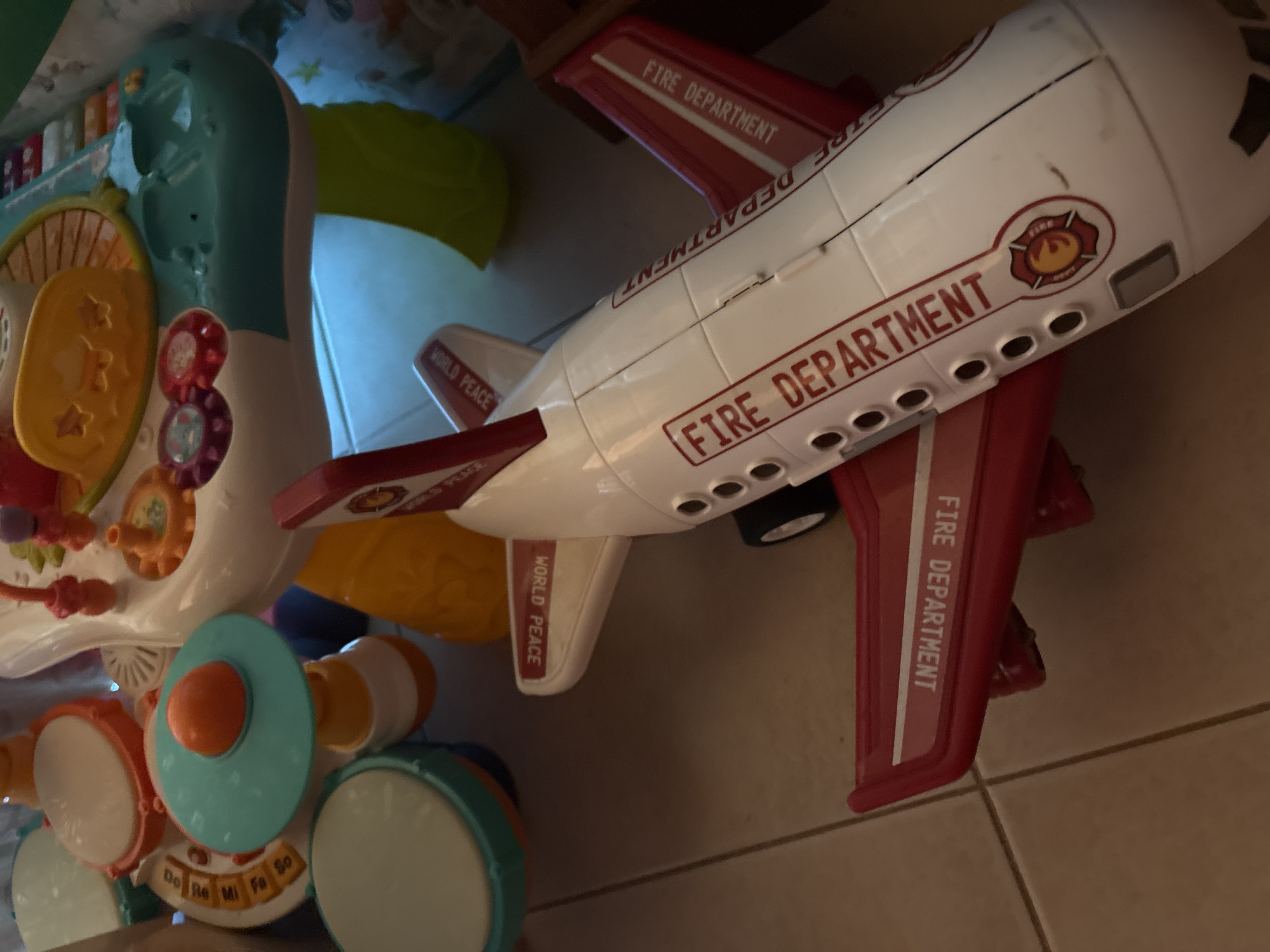 The image shows a close-up of various children's toys. A white and red toy airplane with "FIRE DEPARTMENT" printed on its side and "WORLD PEACE" on its wings dominates the scene. It appears to be made of plastic. Adjacent to the airplane, there is a baby walker or activity center with drums and other interactive elements. The background is blurred, but glimpses of other toys and a tiled floor are visible. The toys are located in บ้านบางรักษ์, Thailand.