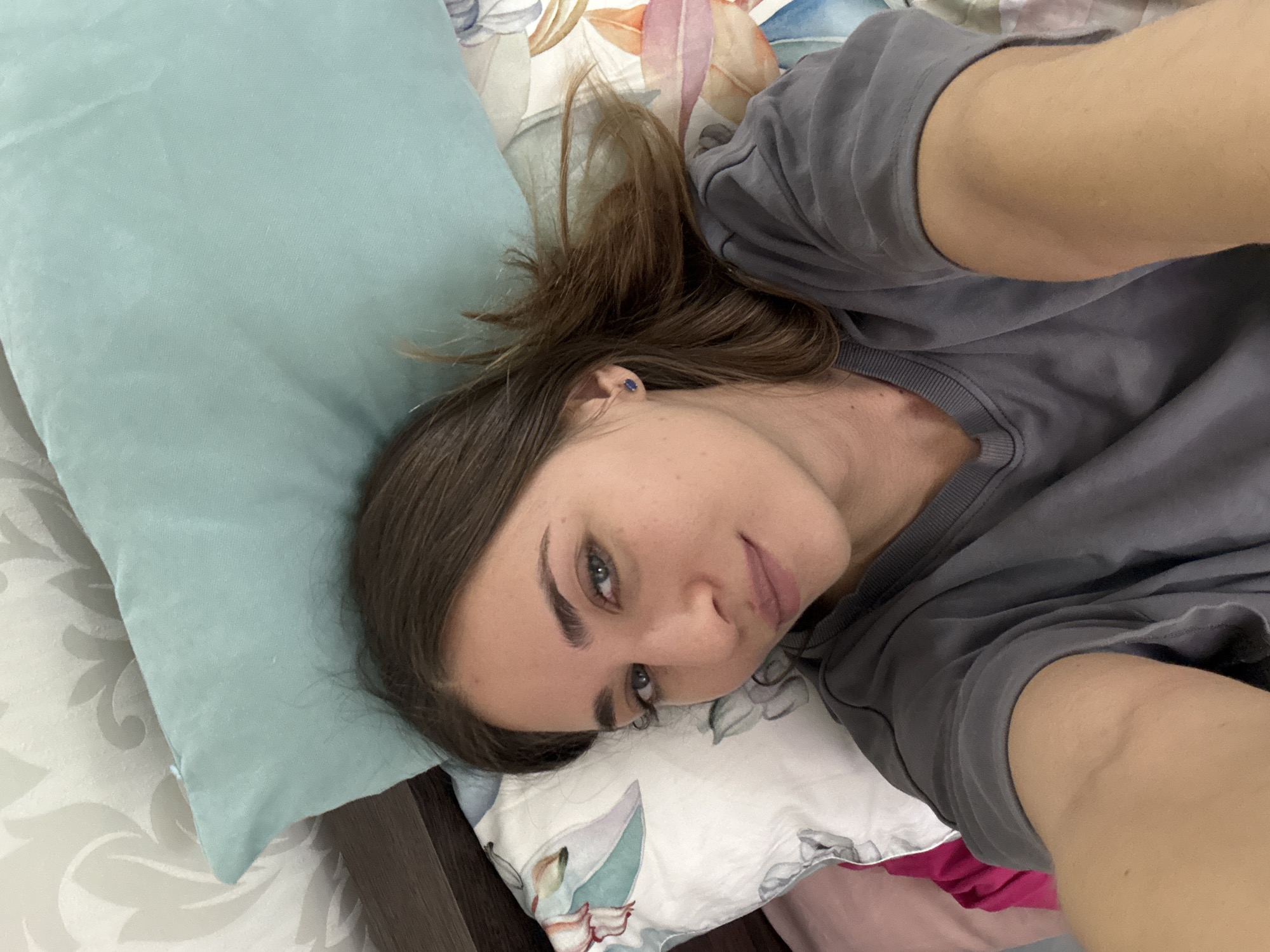A young adult woman is lying on a bed. Her head rests on a light blue pillow. She has brown hair and fair skin. She is wearing a grey shirt. The bed has floral patterned bedding and a white patterned sheet. The image is close-up and taken from a high angle. The location is impossible to discern from the image and the city of Saint Petersburg, Russia cannot be confirmed.