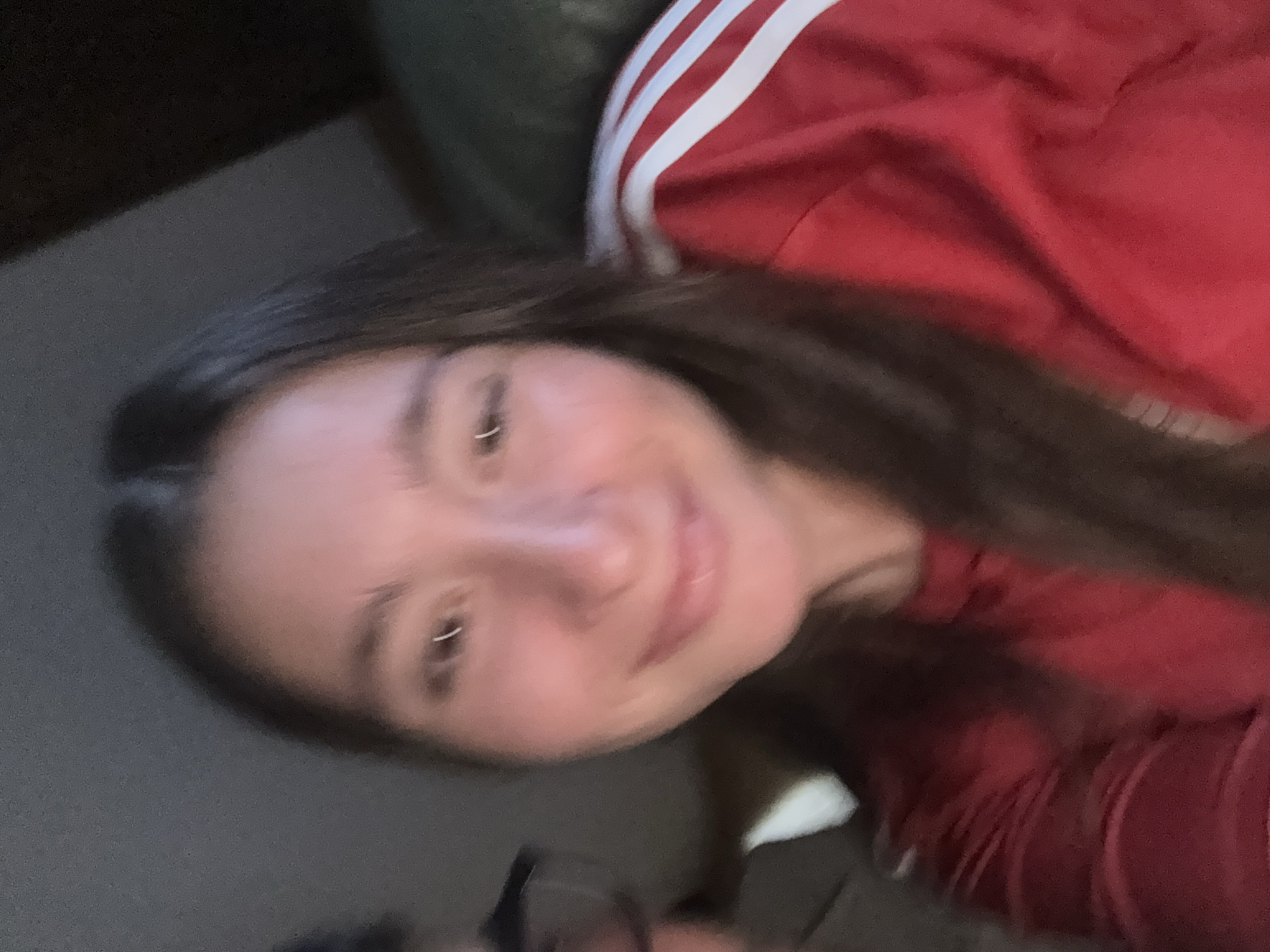 In this close-up, low-light selfie, a young woman with dark hair and a light complexion is the primary subject. She is smiling and looking towards the camera. She is wearing a red shirt with white stripes on the sleeves. To her left, partially visible, is another person wearing glasses. The background is dark and indistinct, suggesting an indoor setting. The image appears to have been taken in Sofia, Bulgaria.