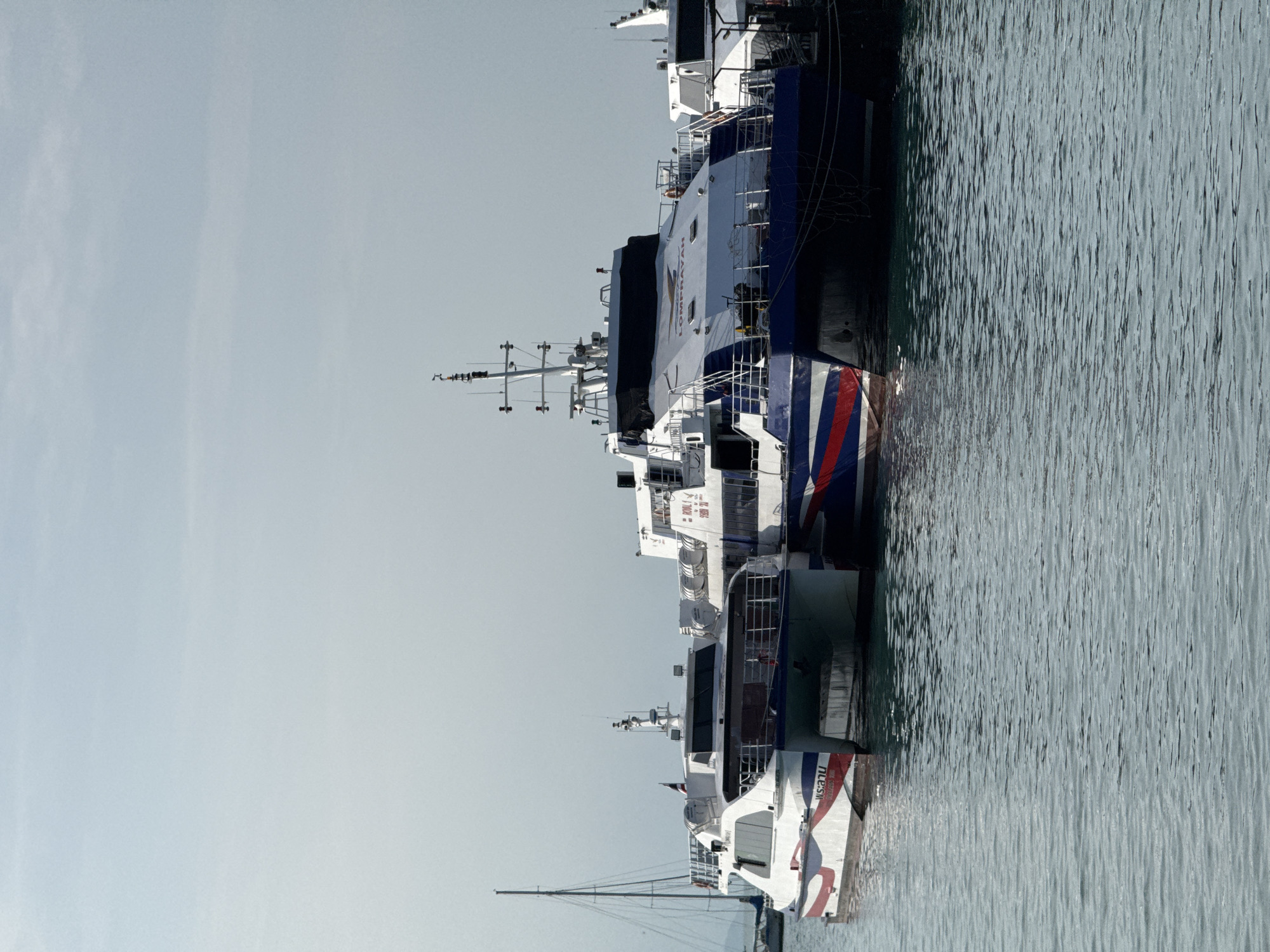 The image depicts a calm, overcast day at a harbor, likely in the Ko Samui District of Thailand, evidenced by the presence of multiple modern catamarans, common for island hopping in such regions. The foreground is dominated by the tranquil, rippling surface of the turquoise sea. Moored side-by-side are two large passenger catamarans. The catamaran on the left is predominantly white with red and blue accents and displays Thai script on its hull. The larger catamaran to its right, with a dark blue hull and red and white stripes along its side, has "LOMPRAYAH" branding prominently displayed on its upper deck. Both vessels appear to be waiting for passengers, with no individuals visible on their decks. In the background, the mast of a sailboat is visible, along with the faint outline of other boats and structures suggesting a bustling port. The sky is a uniform, pale blue, indicating diffused light.
