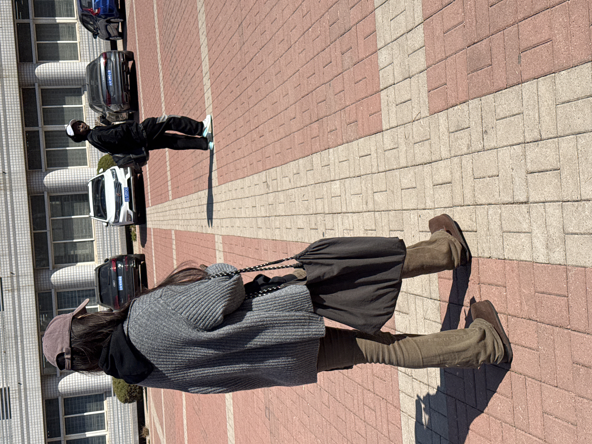 The scene depicts two individuals on a brick-paved area adjacent to a building in Zhongtun, China. One individual, closer to the camera, wears a grey sweater, dark trousers, brown boots, and a baseball cap, carrying a black drawstring bag on their back. They are walking away from the camera. The second individual, positioned further away near parked cars and the building, wears a black jacket, trousers, and a white hat. They are walking in the opposite direction. Several cars are parked nearby, including a white sedan, a dark SUV, and a blue car further in the background. The building has multiple windows and light-colored walls. The overall setting appears to be a paved area or walkway next to a building, possibly a campus or residential area.