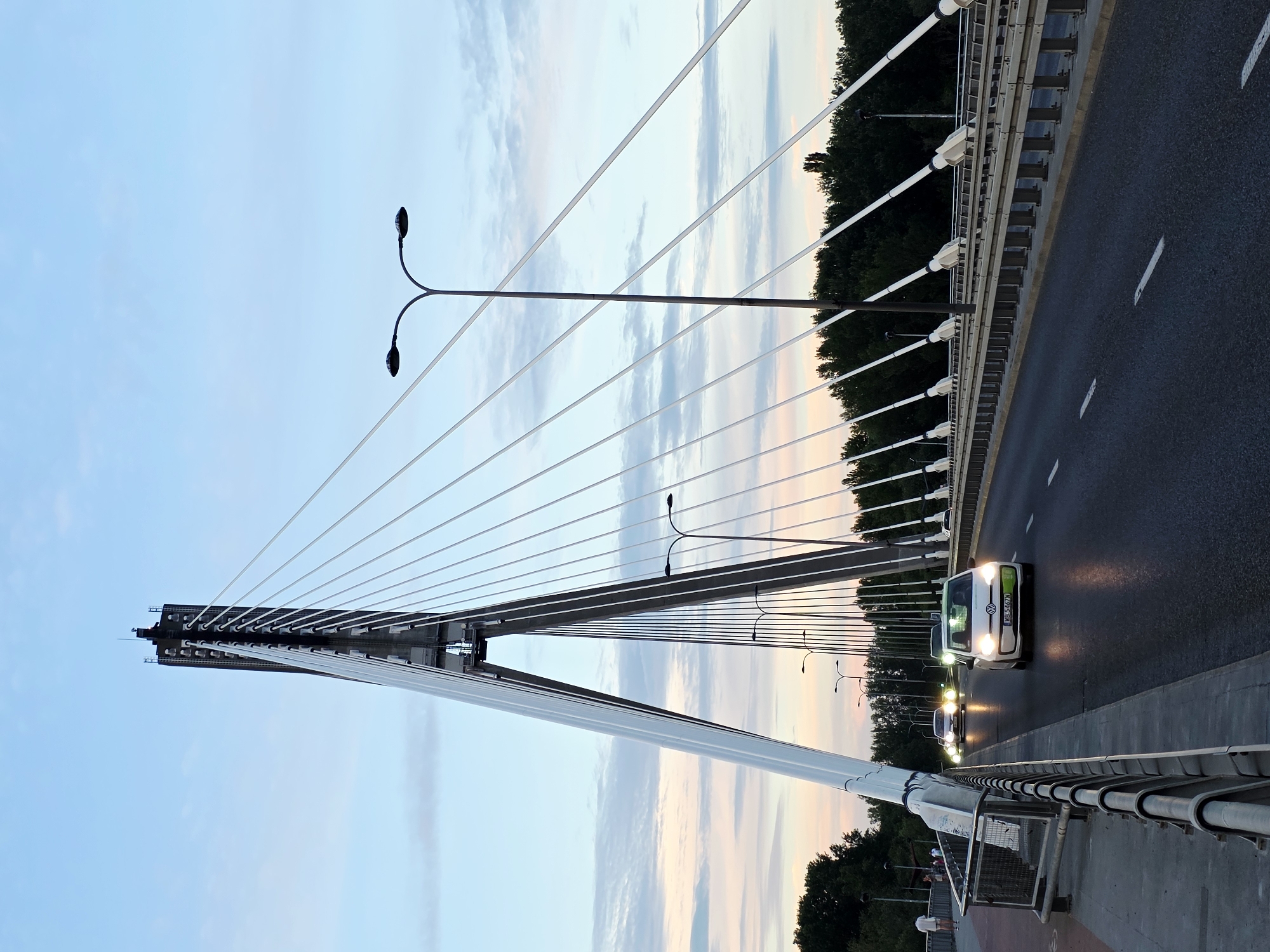This is an eye-level, full shot of the Świętokrzyski Bridge in Warsaw, Poland, captured at dusk. The modern, cable-stayed bridge dominates the frame, with its tall, angular pylon rising towards a pale blue sky streaked with pink and orange hues from the setting sun. Numerous white cables fan out from the pylon, supporting the bridge deck.
The bridge carries traffic, with several cars visible on the roadway. The closest vehicle is a white Volkswagen Up! with its headlights illuminated, and its license plate reading "WH 5467N". Another car is approaching from the distance, its headlights also on. Pedestrians are visible on the left sidewalk, one of whom is walking away from the camera.
Streetlights with a distinctive curved design are mounted on tall poles along the bridge, their lights not yet fully illuminated. The roadway is asphalt with white lane markings. On the far side of the bridge, a dense line of dark green trees forms a backdrop against the sky. A metal railing lines the pedestrian walkway and the edge of the roadway. The overall atmosphere is serene, transitioning from day to night.
