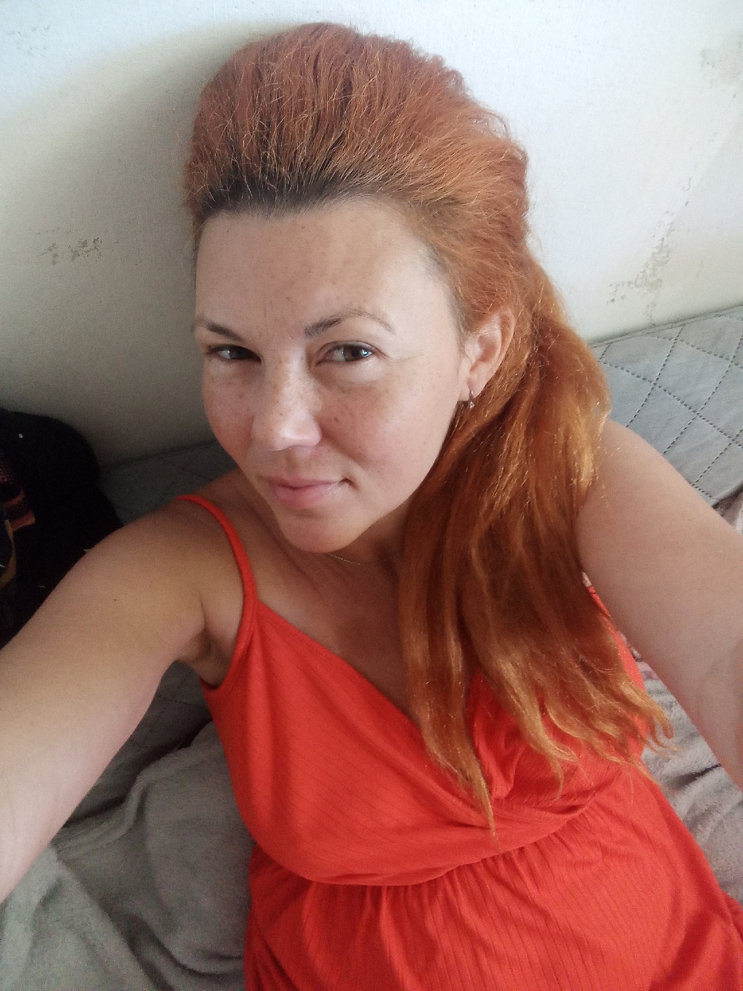 A woman with long, reddish-orange hair is captured in a medium close-up shot, looking directly at the camera with a slight smile. She is wearing a bright orange, strappy dress. Her face is fair-skinned with a smattering of freckles across her nose and cheeks. She has light brown eyes and her hair is styled with volume on top and cascades down her right side. A silver earring is visible in her left ear. The background appears to be a lightly textured, off-white wall and a gray, quilted surface, possibly a sofa or bed. The lighting suggests an indoor setting.