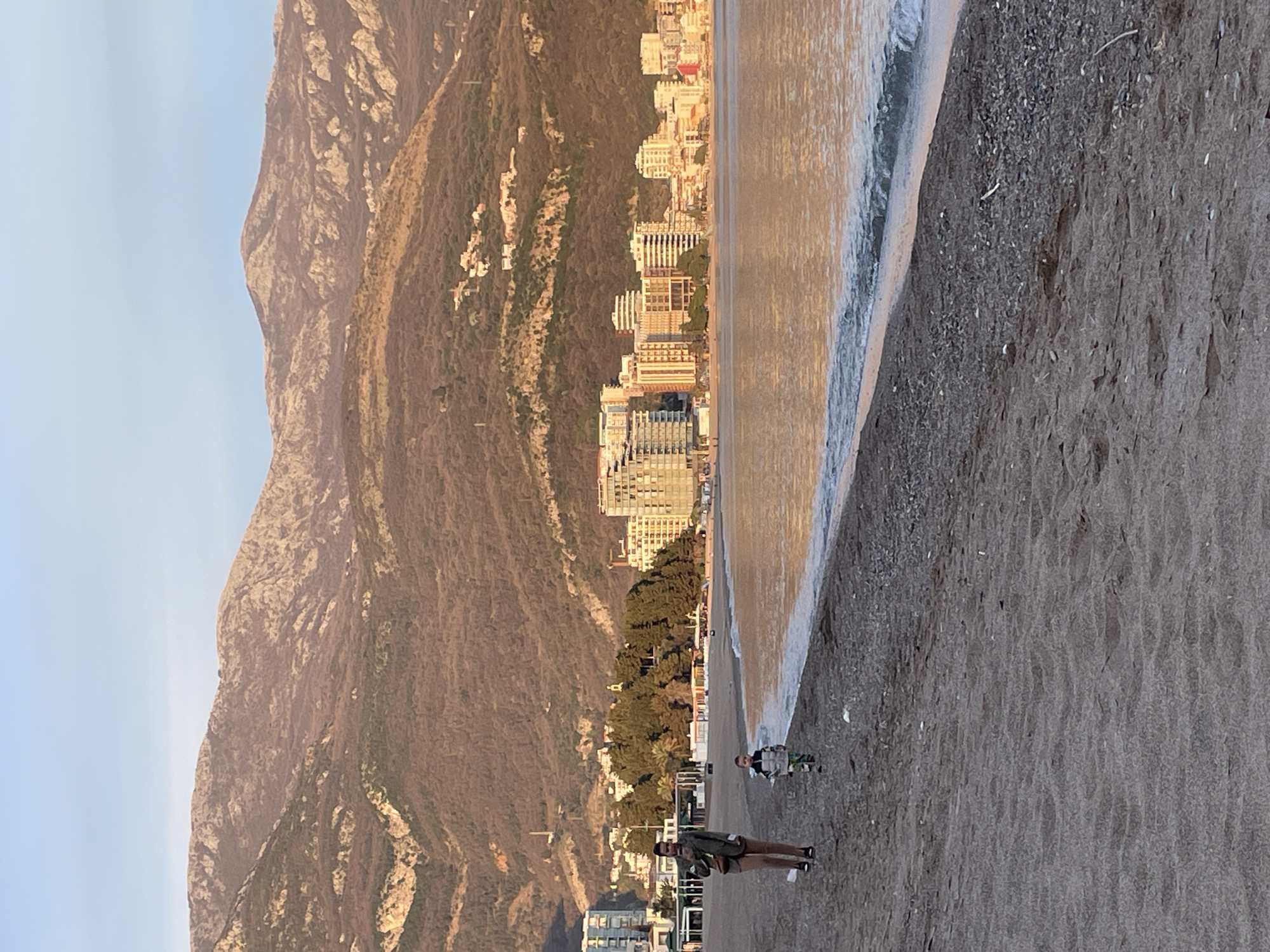 An outdoor scene captured at Bečići, Montenegro, depicts a beach, the Adriatic Sea, coastal buildings, and mountains under a late afternoon or early morning sky.
In the foreground, a sandy beach extends from the left to the right of the frame, transitioning into a strip of small pebbles at the water's edge. Gentle waves are breaking along the shore, reflecting a warm, golden light. Two individuals are walking on the sand and pebble mix. An adult female, estimated to be in her 20s-30s, is positioned left of center, walking away from the camera with her head slightly turned right. She wears a dark jacket, light-colored shorts or skirt, and dark shoes. A shoulder bag is visible on her left side, and she holds a mobile phone in her right hand. Slightly ahead and to her right, closer to the water, walks a small child, approximately 3-5 years old, wearing a multi-colored patterned jacket and trousers. The child is looking towards the water.
The midground features a developed coastline across the bay. Numerous multi-story buildings, primarily modern hotels and apartment complexes, line the shore. These structures display a variety of architectural styles and colors, with lighter facades prominently reflecting the warm sunlight. A section of dense green foliage, including palm trees, is visible between some buildings and closer to the beach, indicating parkland or forested areas.
The background is dominated by large, rugged mountains rising directly behind the coastal development. The mountains are characterized by rocky outcrops and sparse, low-lying vegetation, predominantly brown and green hues. The upper slopes are illuminated by the warm, low-angle sunlight. The sky above the mountains is clear and light blue.