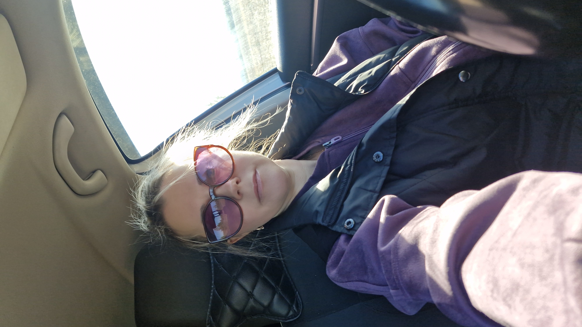 The media file presents a close-up, slightly tilted view of a person inside a car during daytime. The primary subject is a person, likely female, with blonde hair styled with wispy strands, wearing oversized, reflective sunglasses with reddish-purple lenses that obscure their eyes. Their lips are visible, showing a neutral or slightly serene expression.
The person is dressed in layers, consisting of a purple full-zip hooded sweatshirt and a dark (possibly black or navy) puffer vest over it. The clothing appears comfortable and casual.
The setting is clearly the interior of a vehicle, suggested by the beige car interior trim, a visible grab handle attached to the car's ceiling, and a quilted black headrest or seat cover. Through the car window on the left, a very bright, clear sky is visible, indicating a sunny day. A hint of a distant, bright landscape or road can be seen through the glare. The strong natural light illuminates the person's hair and reflects prominently off their sunglasses.
There is no specific activity depicted beyond the person being seated in the car and posing for the photograph. The time of day is clearly daytime, and the weather appears bright and clear. No discernible text is visible in the image.