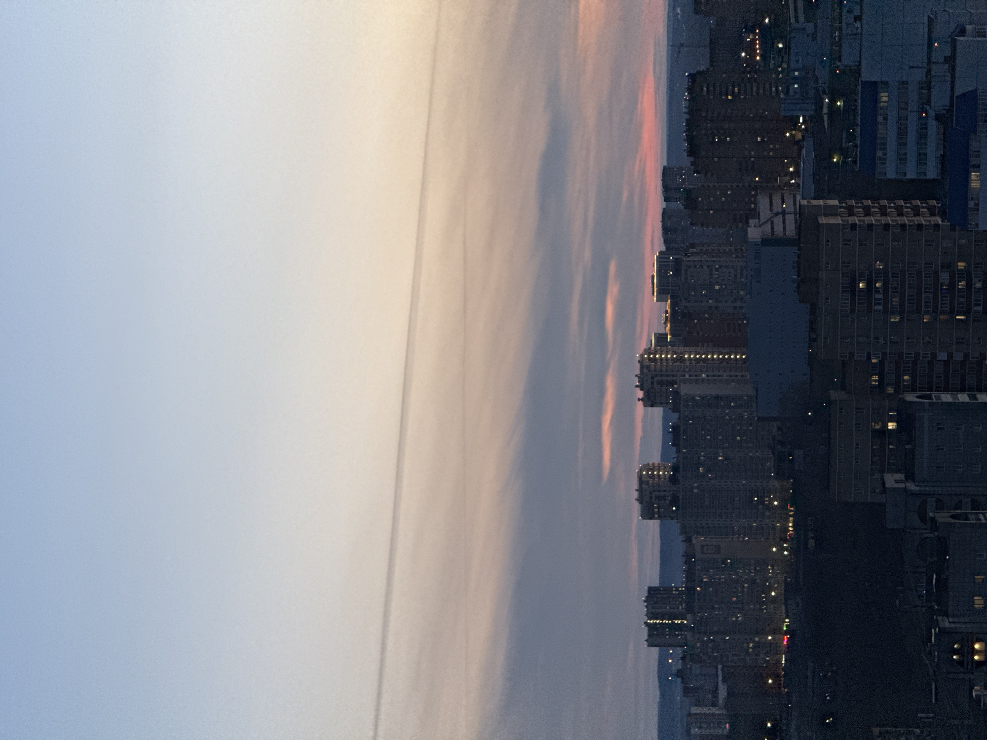 The image displays an urban landscape at either dawn or dusk, viewed from an elevated perspective. The sky occupies roughly the upper two-thirds of the frame, transitioning from a light, clear blue on the left to warmer tones of pale orange, pink, and light yellow towards the right, suggesting the direction of the rising or setting sun. A prominent, long, thin, straight line, likely a contrail from an aircraft, stretches horizontally across the sky, dividing the lighter and warmer hues. Scattered, wispy clouds are also visible, softly reflecting the sky's colors.
The lower third of the image is dominated by a dense cityscape, featuring numerous multi-story buildings, possibly apartment blocks or commercial structures, typical of a large urban center like Perm, Russia. These buildings appear as dark silhouettes against the fading light of the sky. Many windows are illuminated with warm light, creating a scattered pattern of bright points across the dark facades, indicating ongoing activity within the buildings. No specific people or distinct objects are discernible. The overall scene conveys a tranquil moment as the city transitions between day and night, with artificial lights beginning to punctuate the urban fabric. There is no visible text in the image.