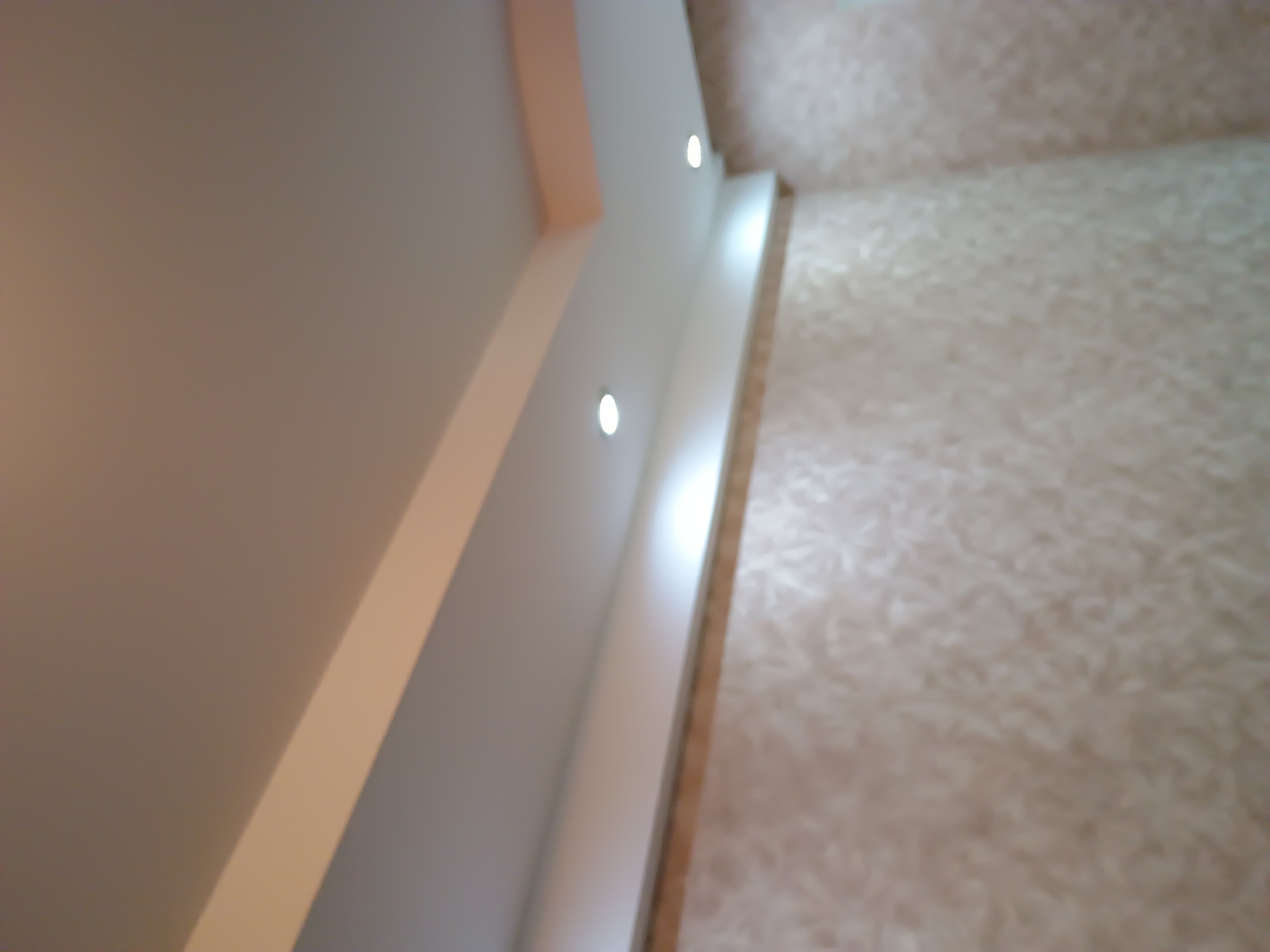 The image shows an interior view of a room, focusing on a multi-level ceiling with recessed lighting. The walls appear to be wallpapered with a subtle, textured pattern in neutral tones. There are two circular light fixtures visible in the ceiling. The overall impression is that of a modern, well-lit interior space. The image does not contain any identifiable landmarks or features that would allow for a specific location within Zaporizhzhia, Ukraine, to be determined.