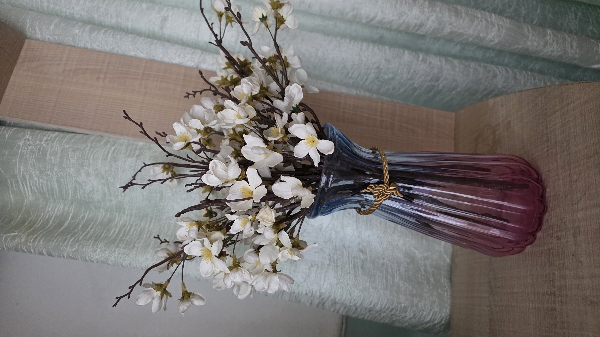 This is a vertical, close-up, eye-level shot of a bouquet of artificial white flowers with brown stems, arranged in a blue and pink gradient glass vase. The vase has a ribbed texture and a decorative gold cord wrapped around its neck. The flowers are clustered densely and extend out from the vase. The background features pale mint green curtains with a textured pattern and a section of a light brown wooden panel. The objects are placed on a light brown wooden surface. There are no people visible in the image. The scene appears to be indoors, possibly in a domestic setting. The location of this image is not specified as Tokmok City, Kyrgyzstan.