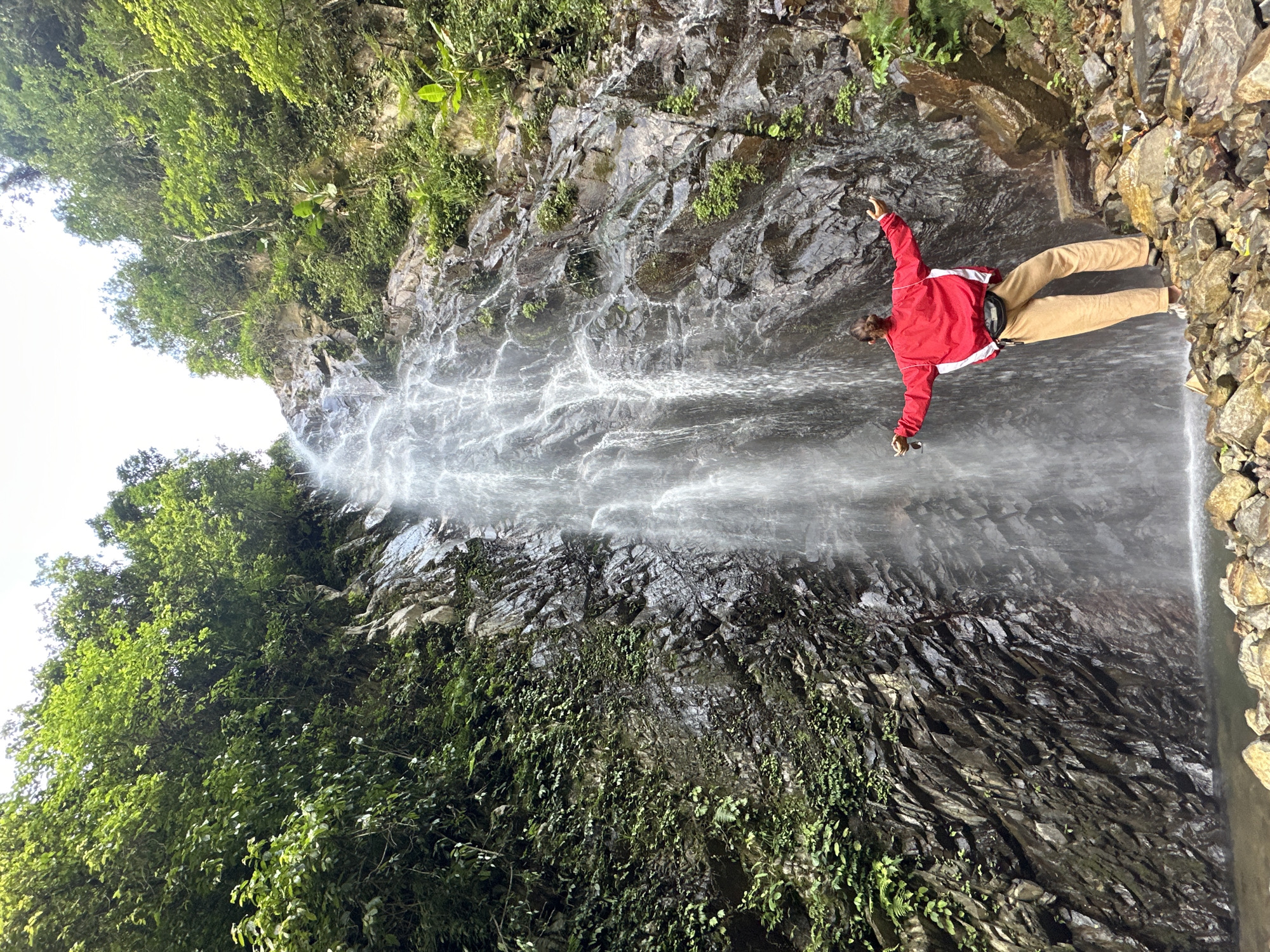 A lone individual stands facing a tall, cascading waterfall in Tu Le, Vietnam. The person, viewed from behind, wears a red and white long-sleeved jacket, light brown pants, and a black fanny pack, with arms outstretched to the sides, holding sunglasses in the left hand. They are positioned on a collection of angular brown and grey rocks at the base of the waterfall. The powerful waterfall descends a steep, dark, and wet rocky cliff face, forming a shallow pool at its foot. The surrounding slopes and the top of the cliff are densely covered with lush, vibrant green vegetation, including various trees and smaller plants. The sky above is bright and appears overcast.