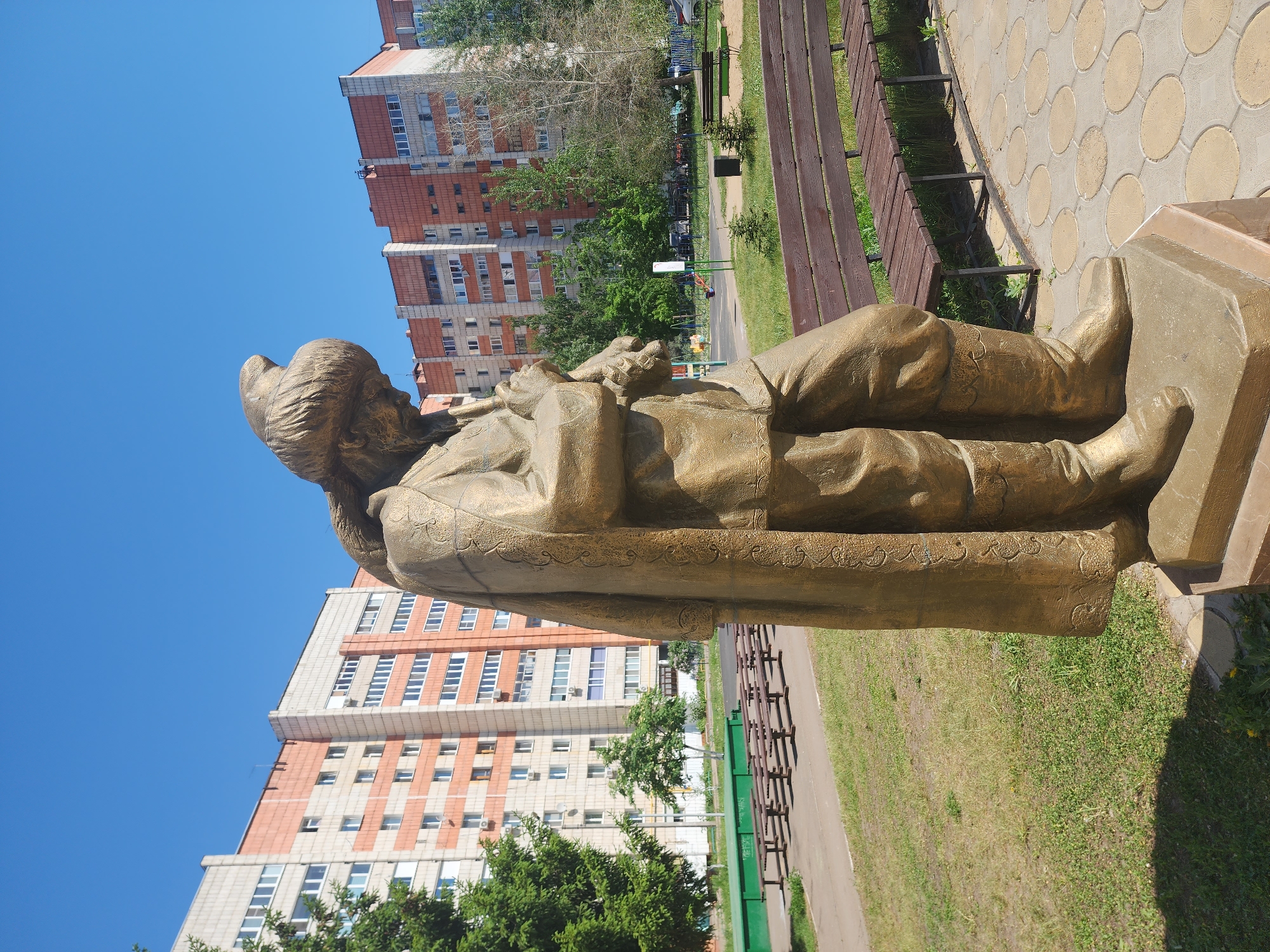 The image shows a full, side view of a bronze statue of a man in Kazan, Russia. The statue depicts a figure with a cap-like head covering, wearing a long robe, with intricate patterns along the lower edge, and holding an object across the body. The statue stands on a square base.
The environment includes a walkway paved with circular stone patterns, bordered by green grassy areas. Benches line the path. In the background are multiple multi-story residential buildings with a combination of light-colored walls and red-brick accents. The sky is a clear blue.