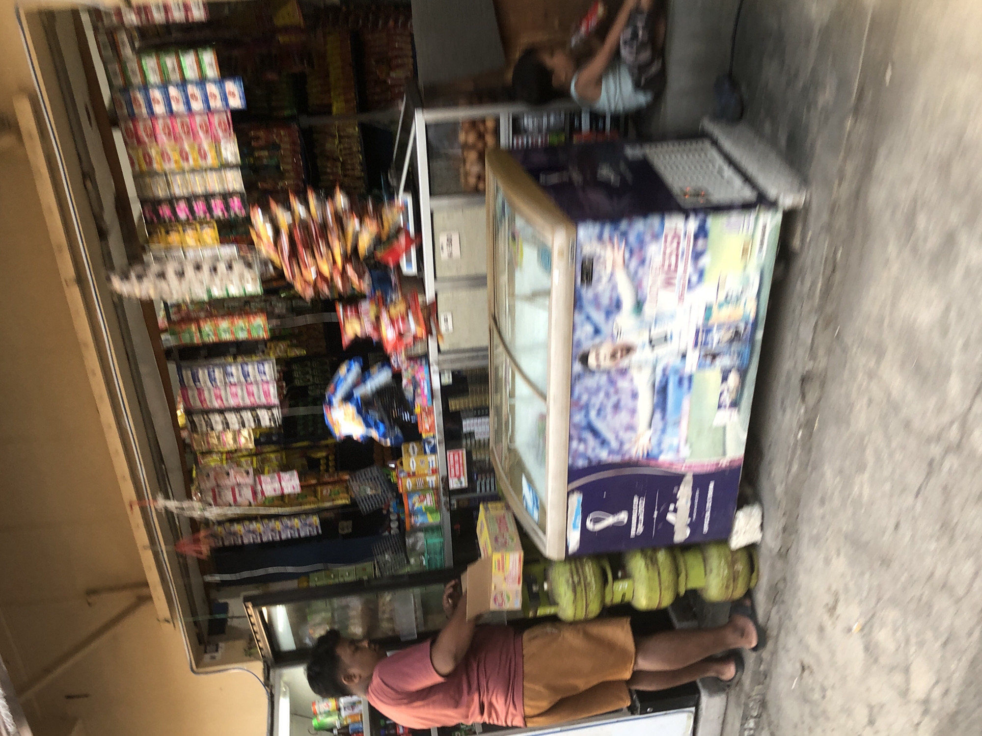 In the city of Tenggulunan, Indonesia, an outdoor convenience store is depicted. A man in a coral-colored t-shirt and orange shorts stands to the left, facing a cooler and interacting with a cardboard box. He appears to be in the process of stocking or organizing items. In front of him, stacked vertically, are several green propane tanks. To his right, a chest freezer, adorned with a graphic of a soccer player, is positioned. A young child, wearing a light blue tank top and patterned shorts, sits on the ground to the right of the freezer, facing away from the viewer. The store's interior, visible in the background, is lined with shelves stocked with various packaged goods, predominantly snacks and drinks. The overall scene captures a moment of commerce and everyday life on a street in Tenggulunan.