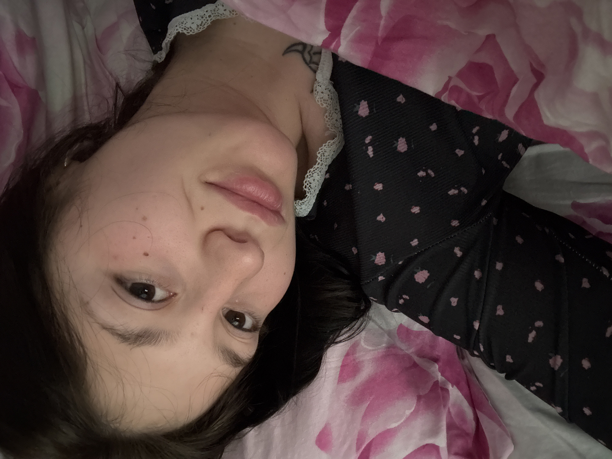 A young woman lies on a bed with a floral patterned blanket. She is wearing a black top with small pink floral patterns and a white lace collar. She has dark hair and light skin with some freckles. A tattoo is visible on her collarbone. Her expression is relaxed. The location cannot be determined from the image.