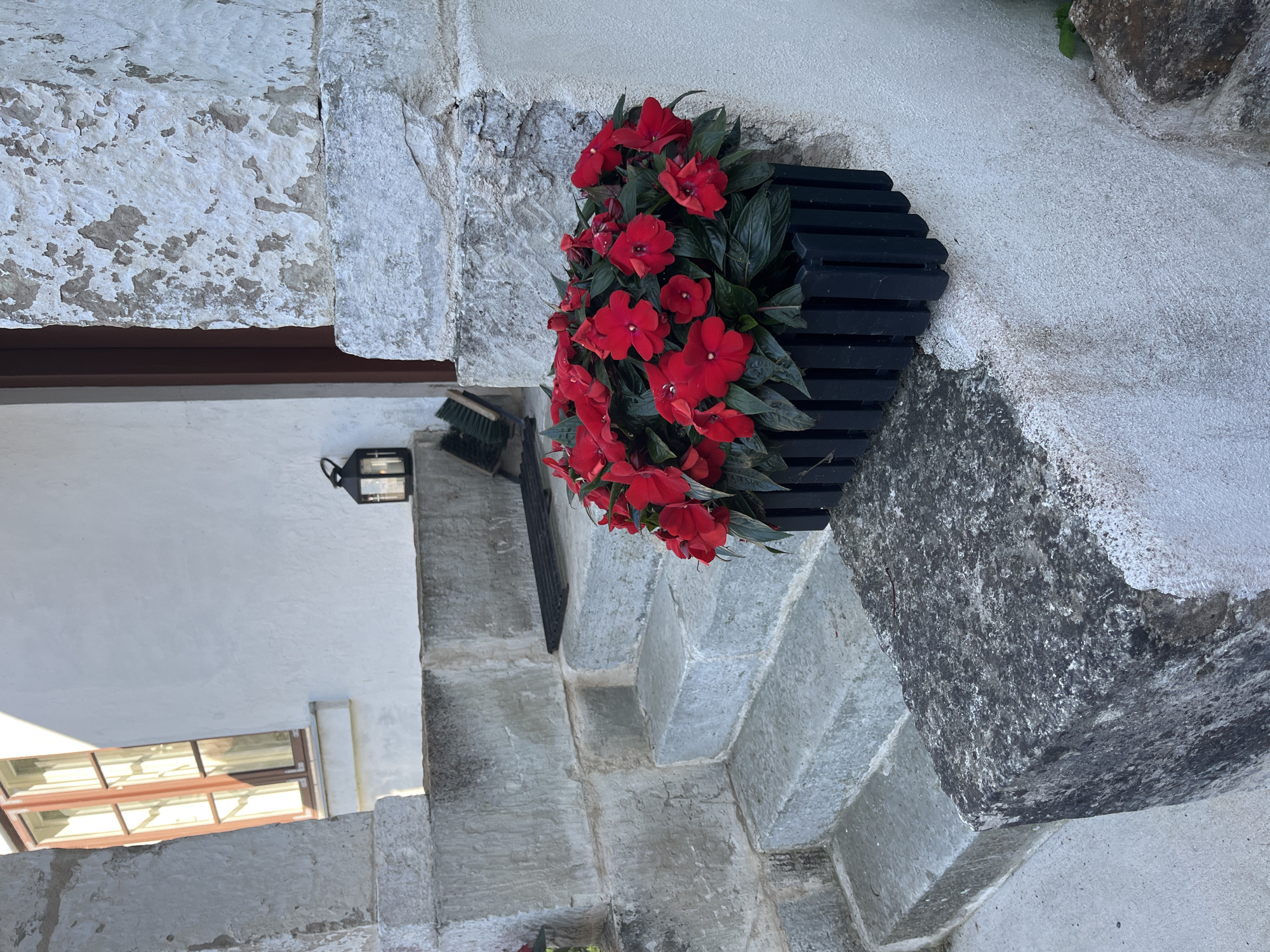 The image depicts the exterior of a stone building with white, weathered walls and a prominent stone staircase. On the right side of the staircase, a black planter box is filled with vibrant red impatiens flowers. A dark metal lantern is visible on a step in the background, along with a broom. The scene suggests a residential or historical setting, possibly in Kuressaare, Estonia, given the architectural style. The lighting indicates it is daytime, with sunlight casting shadows.