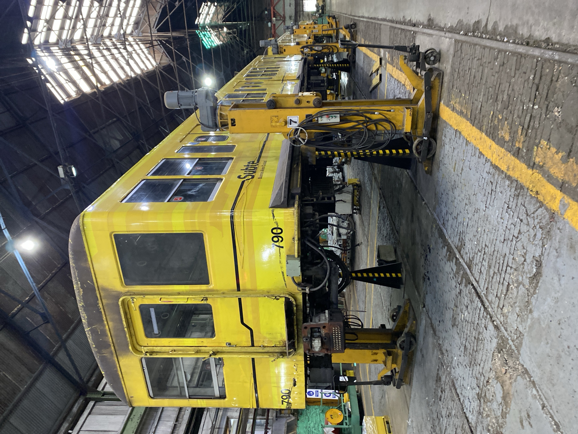 A yellow "Subte" (Buenos Aires Subway) railcar, numbered 790, is situated inside a large, industrial maintenance depot. The railcar is elevated on yellow maintenance equipment. The depot's interior has a high ceiling with exposed metal beams and skylights, and the floor is concrete with marked yellow safety lines.