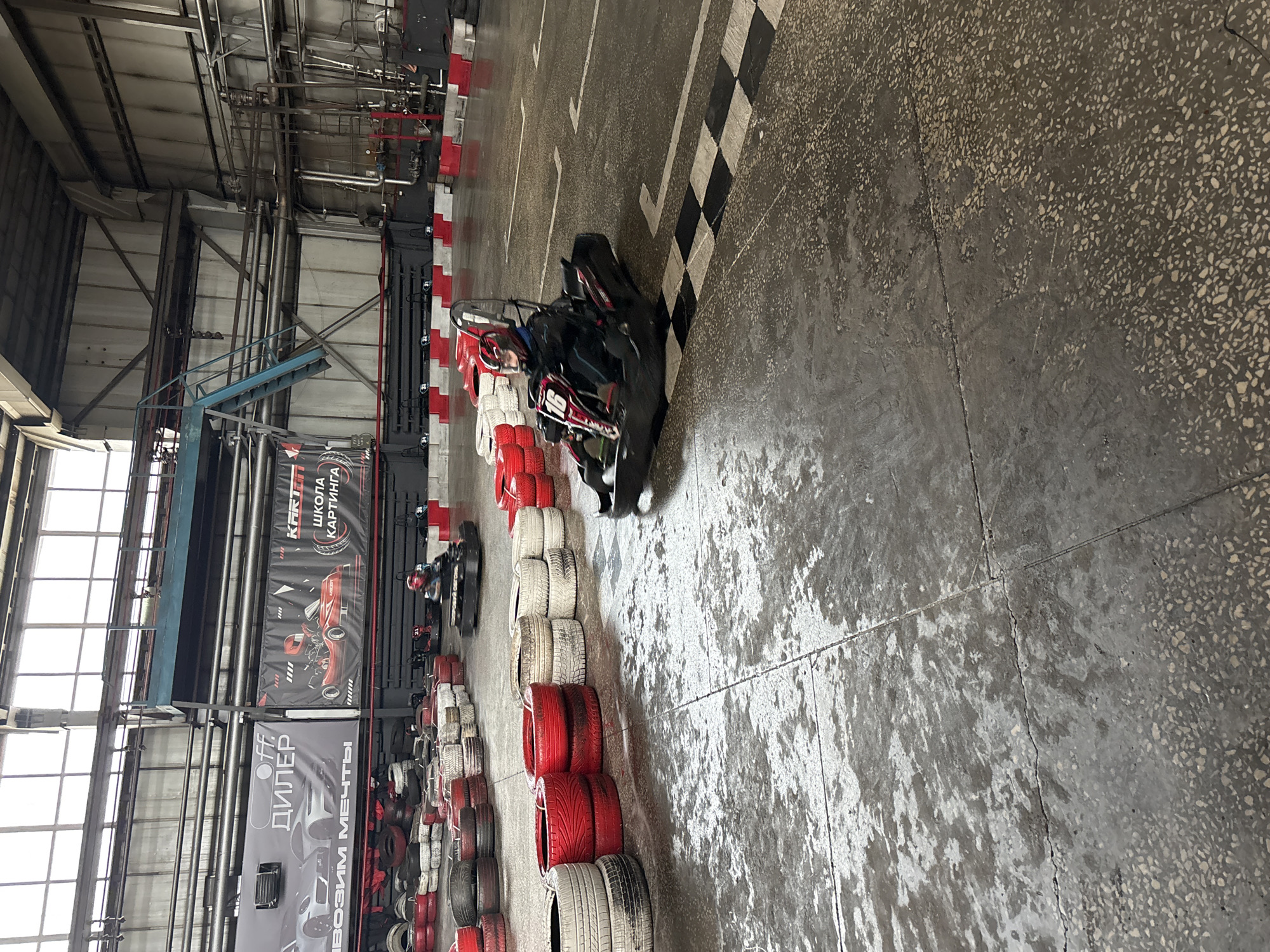The image depicts an indoor go-karting track located in Perm, Russia. A black go-kart is positioned on the track, oriented towards the right side of the frame. The floor is concrete, marked with a black and white checkered line. Tires painted red and white are stacked along the left side of the track, serving as barriers. In the background, banners with Russian text and images of cars are visible. The building's structure is industrial, with exposed metal beams and pipes overhead. Windows are present on the left, providing natural light.