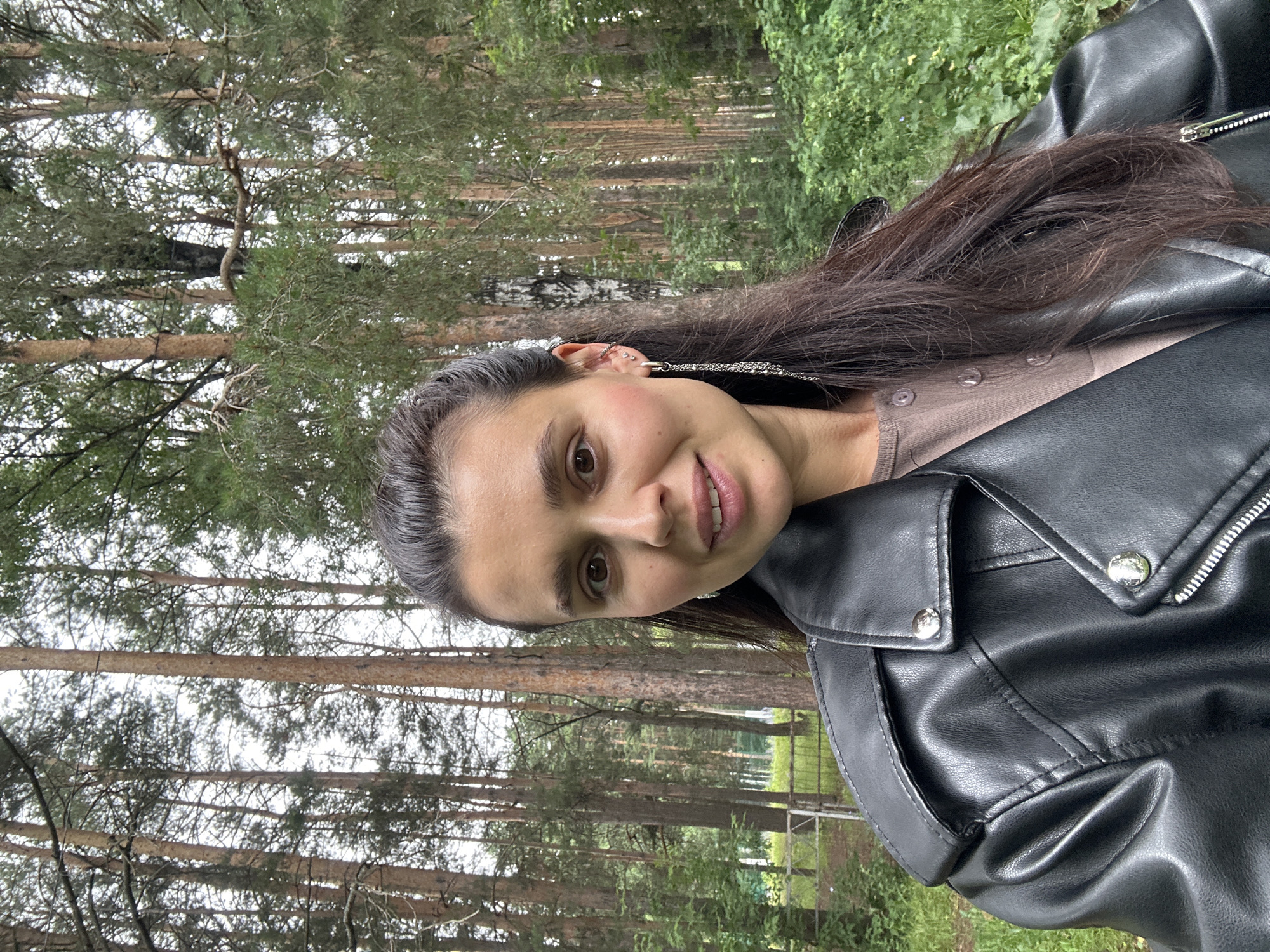 This is a portrait of a young woman in Chaikovsky, Russia. She has long, dark brown hair and is wearing a black leather jacket over a taupe, collared shirt. The woman is smiling slightly and looking towards the camera. She has multiple piercings in her left ear and a delicate necklace.
The setting appears to be a park or wooded area, with tall pine trees and lush green foliage visible in the background. The lighting suggests it might be daytime, possibly overcast, as there are no strong shadows. The woman's posture and expression convey a casual and relaxed mood. The image is oriented in landscape format.