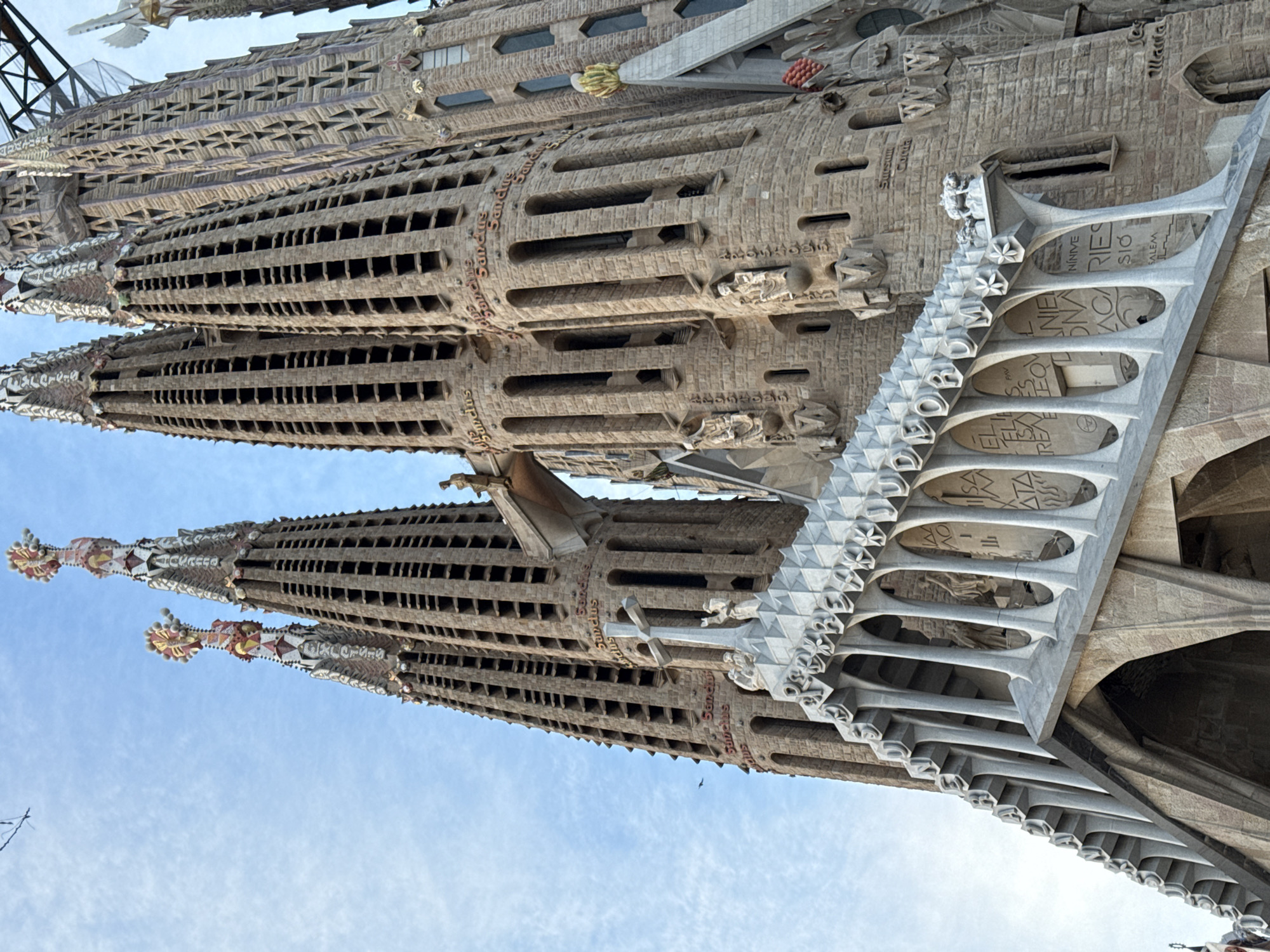 The image displays an upward-angled, detailed view of the Sagrada Familia basilica located in Barcelona, Spain. The scene is dominated by multiple tall, conical stone spires constructed from light brown masonry, featuring numerous rectangular openings along their vertical extent. These spires culminate in intricate, colorful mosaic finials, adorned with spherical elements and crosses. Inscriptions such as "Sanctus" are visible in raised relief around the circumference of several spires. A stone bridge-like structure connects two central spires at an upper level, supporting a small human-like statue. A spire to the far right exhibits a distinct geometric, triangular pattern in its stonework, with structural scaffolding or support beams partially visible against the sky behind it. Below the spires, a complex façade includes a prominent white/light gray stone balustrade with ornate, sculpted vertical supports and horizontal elements bearing raised Latin inscriptions, including parts of "DEORUM." Various stone figures, including angels and a large white cross, are integrated into the lower architectural features. The sky is light blue with scattered, wispy white clouds. A thin, dark linear object is present in the upper left sky, and a few small, dark bird-like shapes are visible near the spires. No living individuals are present in the frame.