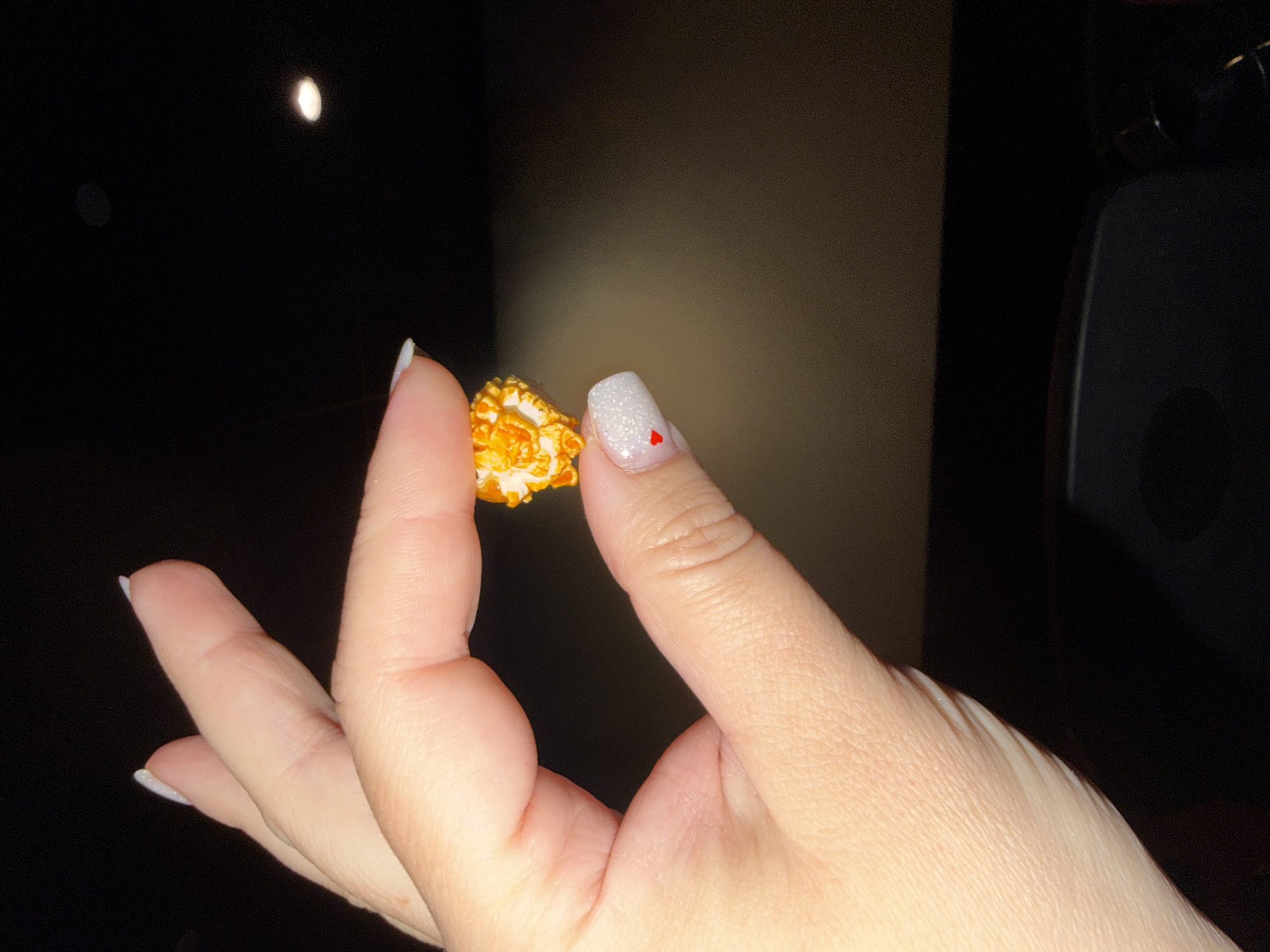 A close-up shot shows a hand holding a piece of caramel corn. The fingernails are white and glittery, and there is a small red heart painted on the nail of the index finger. The background is dark, and there is a light source that appears as a small orb in the upper left corner of the image. A dark object, possibly part of furniture or equipment, is visible on the right side of the frame.