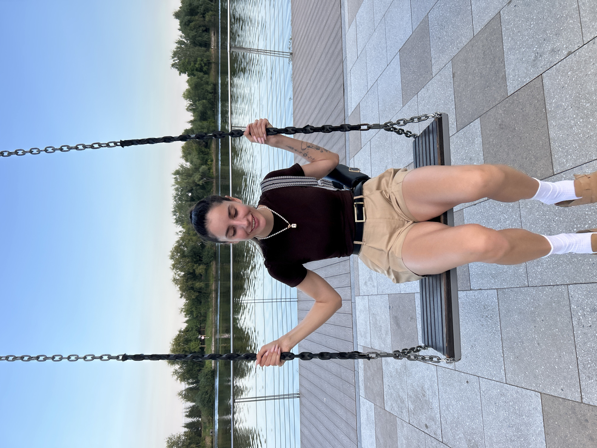 A young woman with dark hair pulled back in a ponytail is sitting on a swing outdoors. She is wearing a dark brown short-sleeved shirt, beige shorts with a black belt, white socks, and tan sandals. She has a necklace with a pendant and a bag slung over her shoulder. She is smiling and looking down. The swing is suspended by chains over a paved area. In the background, there is a body of water with a shoreline lined with trees. The sky is clear and blue. The location appears to be a park or waterfront area in Moscow, Russia, likely during the daytime given the lighting.