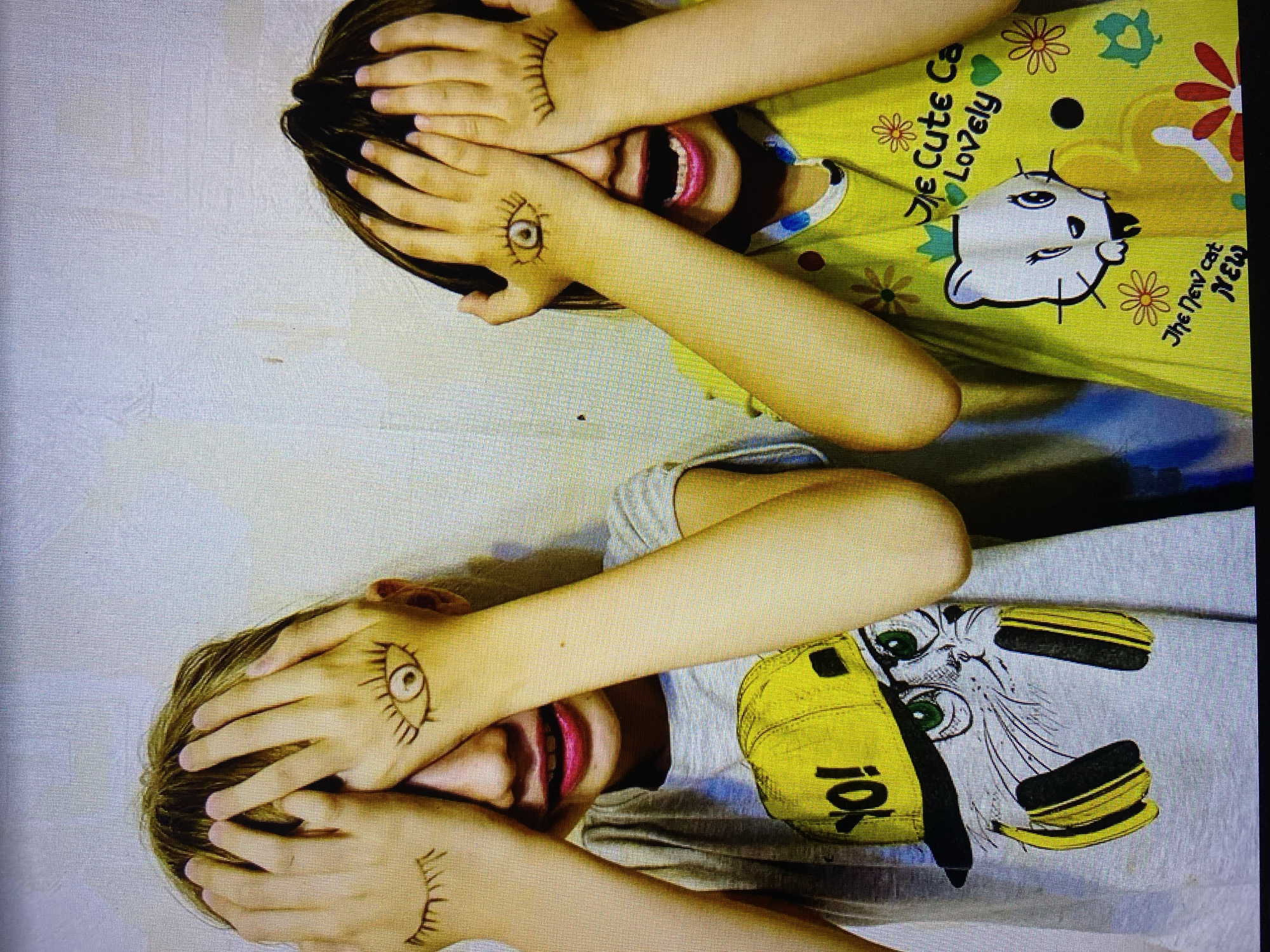 The image depicts two children, their faces partially obscured by their hands, which are decorated with drawn eyes. The child on the left wears a grey t-shirt featuring a graphic of a cat wearing headphones and a yellow cap with "YO!" written on it. The child on the right wears a yellow t-shirt adorned with cartoon cats, flowers, and the text "The Cute Cat Lovely The New Cat NEW". Both children appear to be in a playful or surprised pose, with their mouths open. The background is a plain, light-colored wall. There are no discernible landmarks or contextual elements that would indicate the location within the city of Stavropol, Russia.