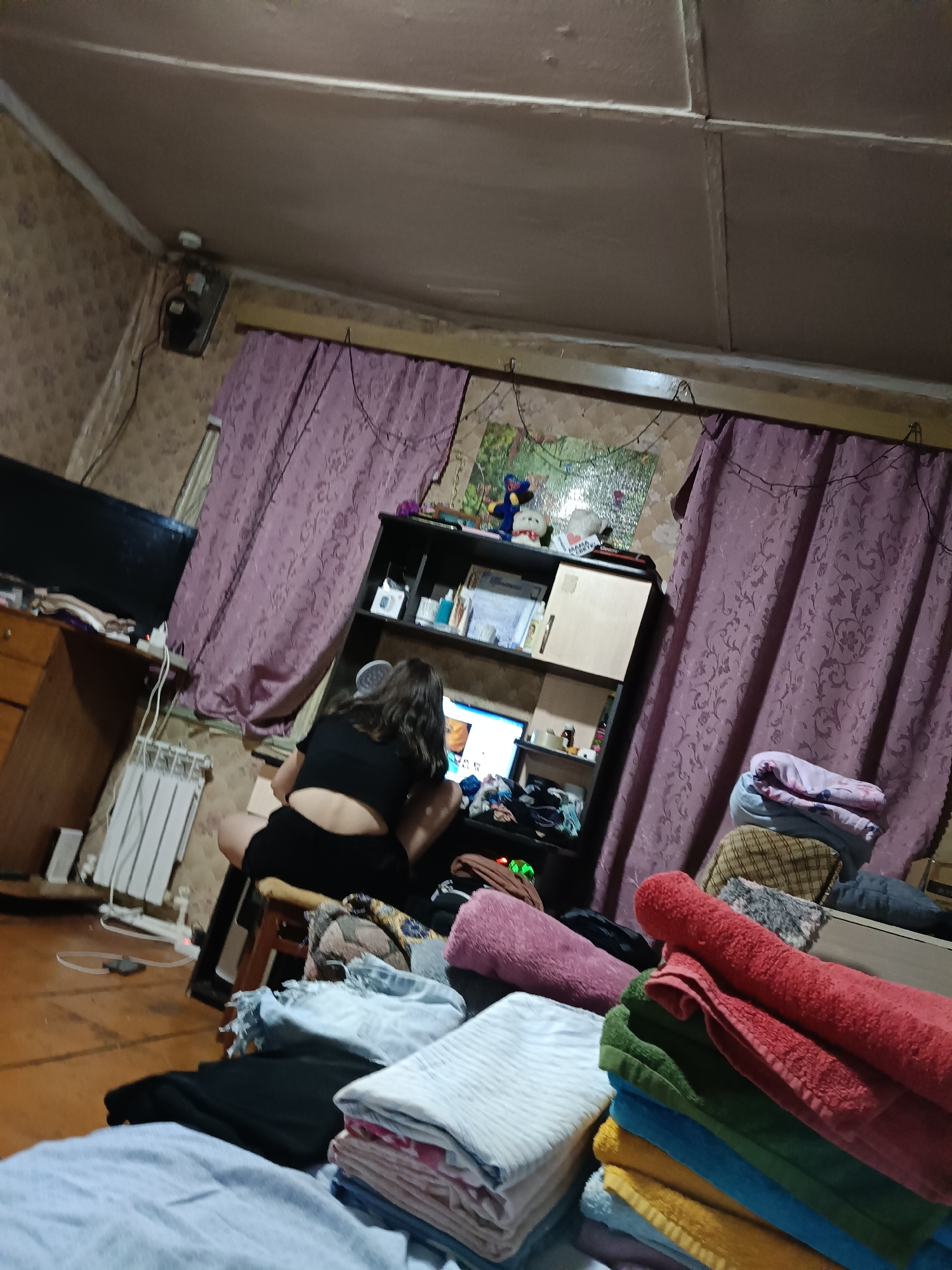 The image depicts an indoor scene, likely a bedroom or living area, with a person sitting at a desk in front of a computer. The room appears cluttered, with items like folded towels, clothes, and various objects on shelves. The person, seen from the back, is wearing a black top and shorts and is focused on the computer screen. The room has patterned wallpaper, purple curtains, and a radiator. The overall impression is of a lived-in space. The location is identified as Evsino, Russia.