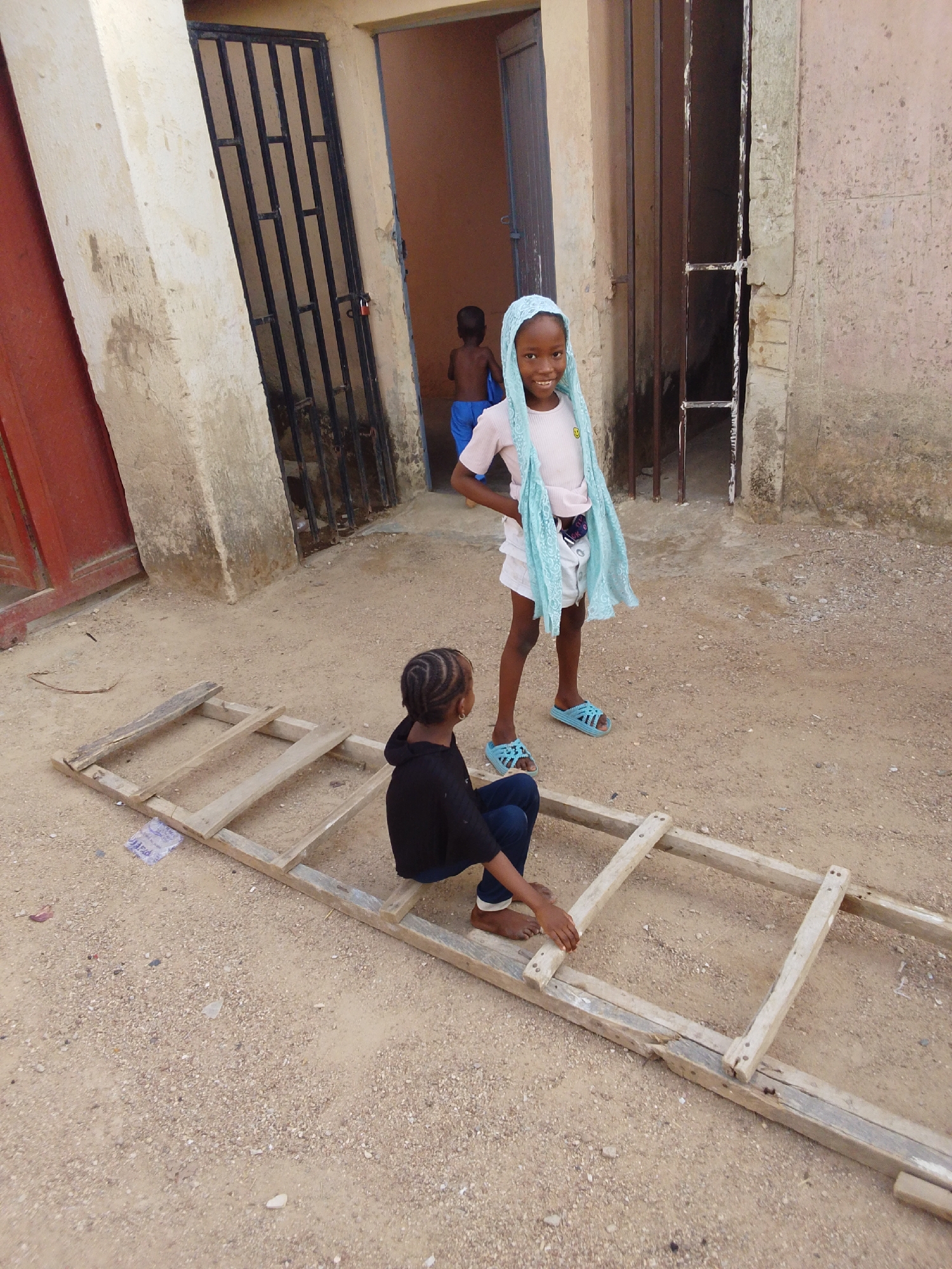 In Bauchi, Nigeria, two children are the focal point. One girl stands with a blue headscarf and matching slides, while the other is seated on a wooden ladder. A third, partially visible child is in the background. The setting features weathered buildings with barred doors and a dusty, gravel ground.