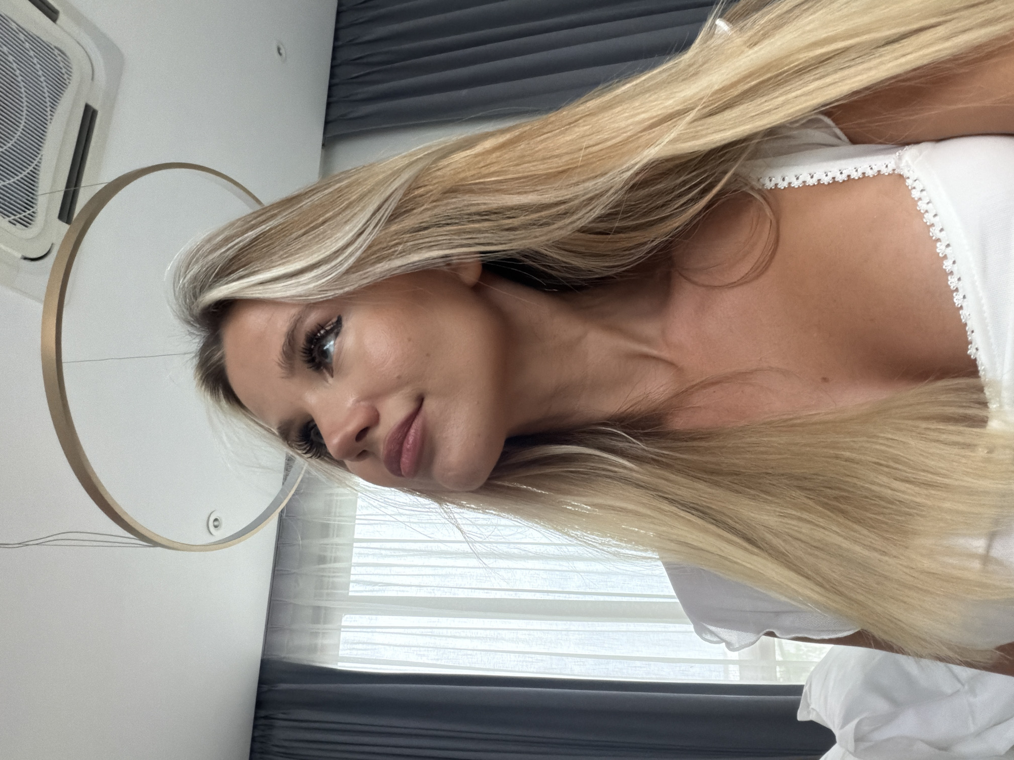 A medium-shot, indoor photograph shows a young, fair-skinned woman with long, blonde hair looking upwards, her head tilted to the left. She is wearing a white top with delicate lace trim on the straps. Her makeup includes dark eyeliner and mascara, and her lips are glossed. The background features a modern ceiling with a circular light fixture and an air conditioning unit. Dark gray curtains are visible on the right, and sheer white curtains hang behind her, suggesting a window. To the left, a white pillow is partially visible. The scene appears to be a bedroom or hotel room.