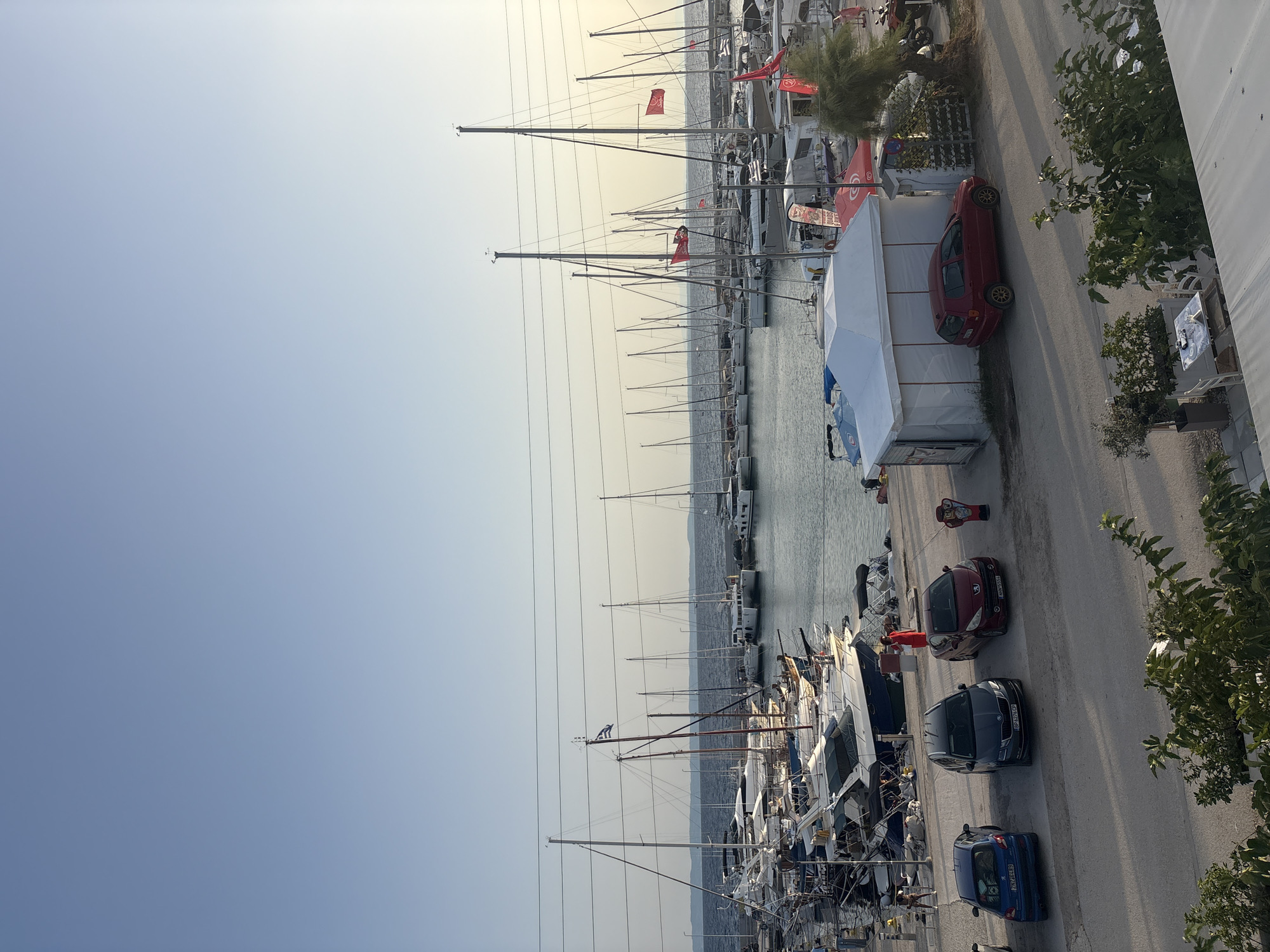 The image shows a marina in Sithonia Municipal Unit, Greece, with numerous sailboats and yachts docked. The sky is clear and blue. In the foreground, there is a paved area with several cars parked, including a blue Peugeot, a dark gray Skoda, and a red Peugeot. A white tent structure is also visible, with a red car parked next to it. A person in a red outfit stands near one of the cars. The water in the marina is calm, and several flags are visible on the boats, including a Greek flag. The scene appears to be in the late afternoon or early evening, with long shadows cast by the boats and vehicles. Lush green foliage is present in the bottom portion of the image.