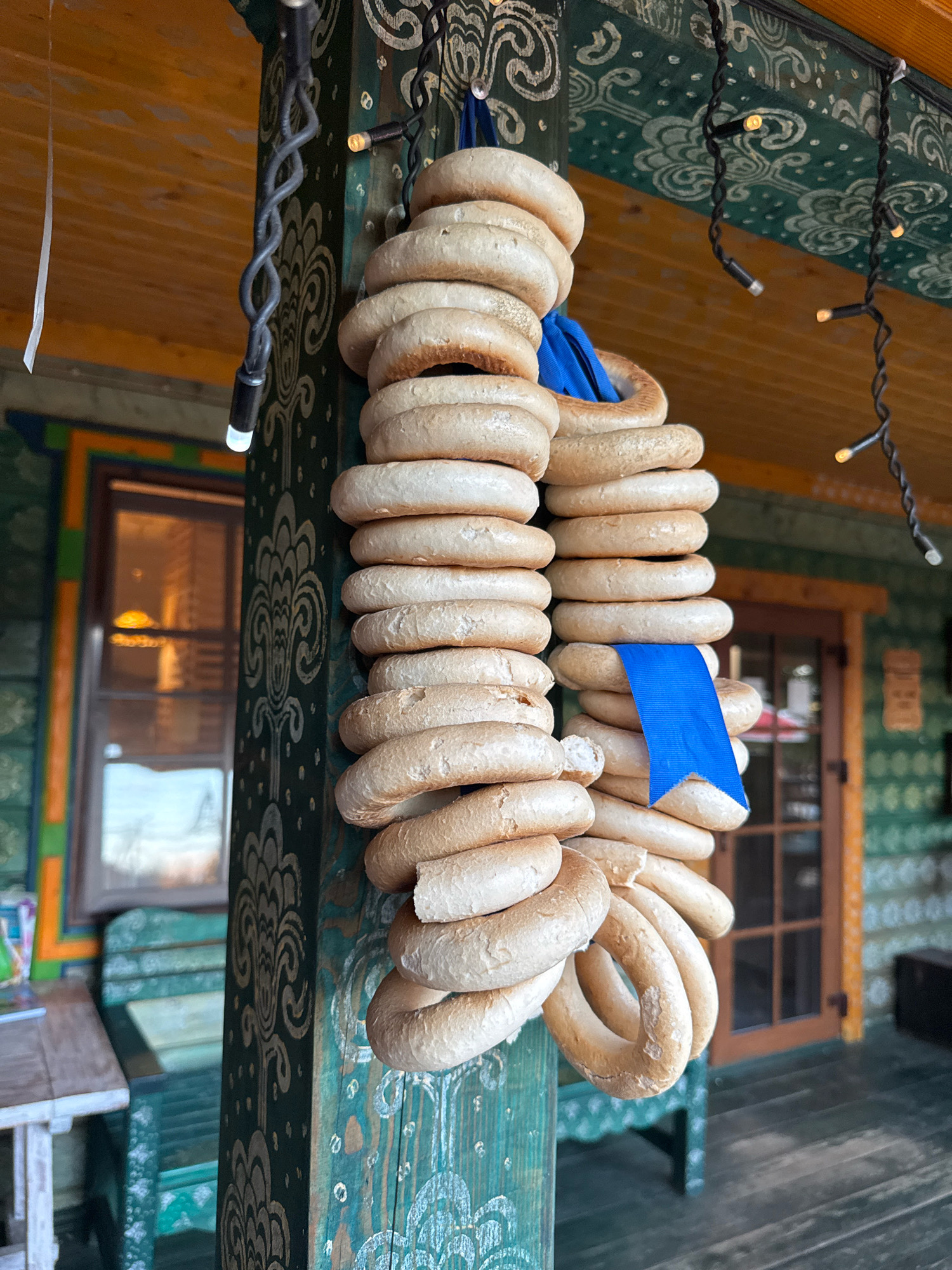 In Valday, Russia, a string of dried bread rings, known as "baranki," hangs from a decorative wooden pillar. The pillar is painted green with white folk art patterns. The baranki are golden brown and appear dry and slightly rough in texture, with some showing darker, more toasted edges. A bright blue ribbon is tied around one section of the baranki. In the background, a wooden building with windows and a covered porch is visible, also decorated with folk art. String lights with small bulbs are draped overhead. A wooden bench and a table with some items on it can be seen on the left side of the frame. The overall scene suggests an outdoor market or a traditional Russian dwelling.