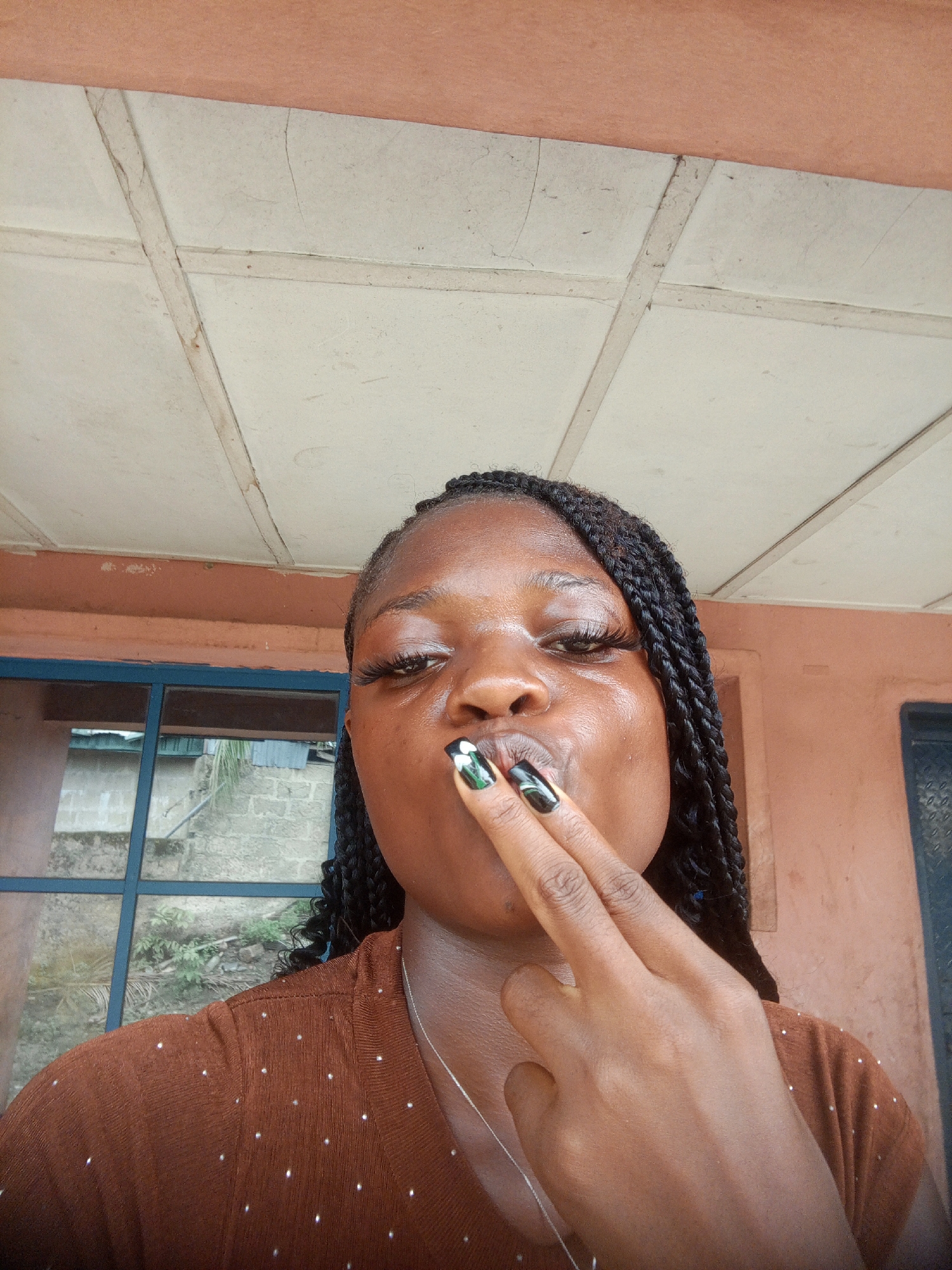 A close-up, low-angle shot depicts a young, dark-skinned woman with braided hair, positioned indoors against a backdrop that suggests a building exterior. Her brown top has small, possibly reflective embellishments. She is holding two fingers to her lips. The fingernails are manicured with a decorative design. Behind her, a window shows an outside view, appearing to be Uyo, Nigeria. The building's exterior wall is a muted terracotta hue. Overhead is a ceiling composed of tiled or paneled sections.