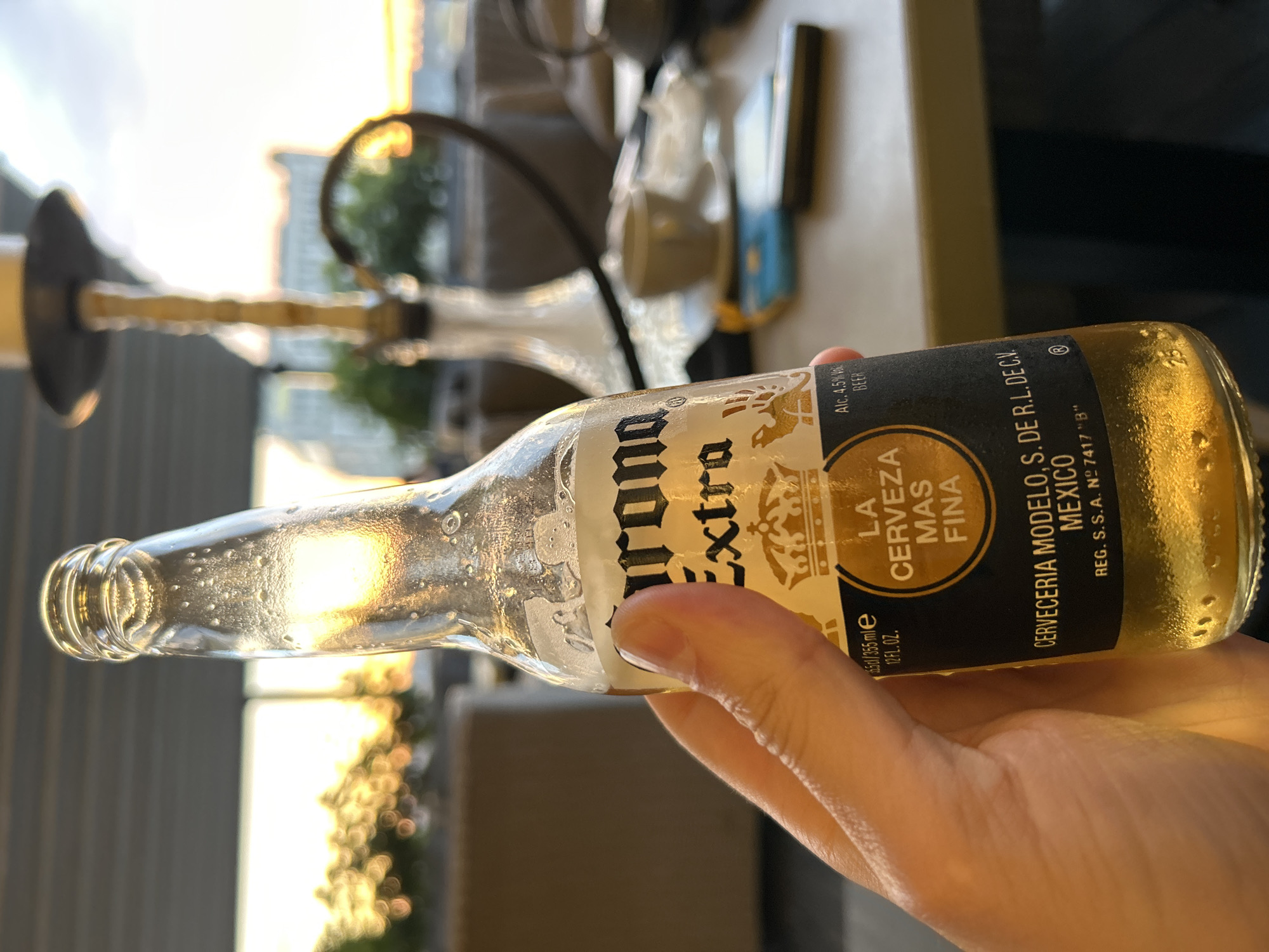A hand holds a cold, sweating bottle of Corona Extra beer. The bottle has a yellow label with the brand name in black. The background is blurred, showing a hookah and a table with some objects, possibly in an outdoor seating area of a restaurant or cafe. The setting appears to be during sunset, with warm light filtering through. There is no indication of the location being Novokuybyshevsk, Russia.