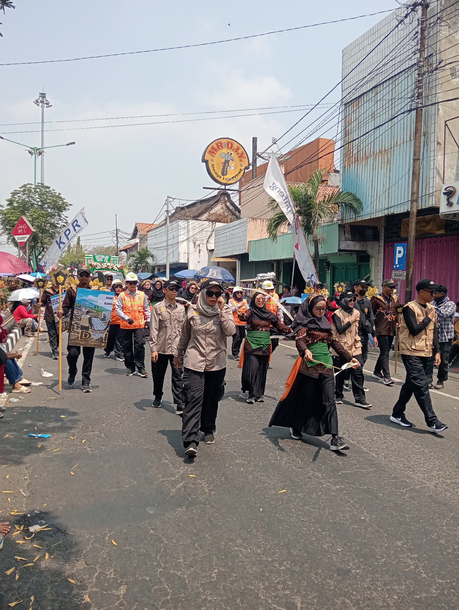 In Demak, Indonesia, a group of people are participating in a parade. The parade is taking place on a paved street lined with buildings. Several participants are wearing uniforms, including safety vests and matching shirts. One person in the front is holding a large sign with an image depicting a landscape. Other participants are carrying flags, including one with "KPR" written on it. Spectators are visible on the side of the street, some sitting and others standing. The weather appears to be clear and sunny. The scene suggests a community event or celebration.