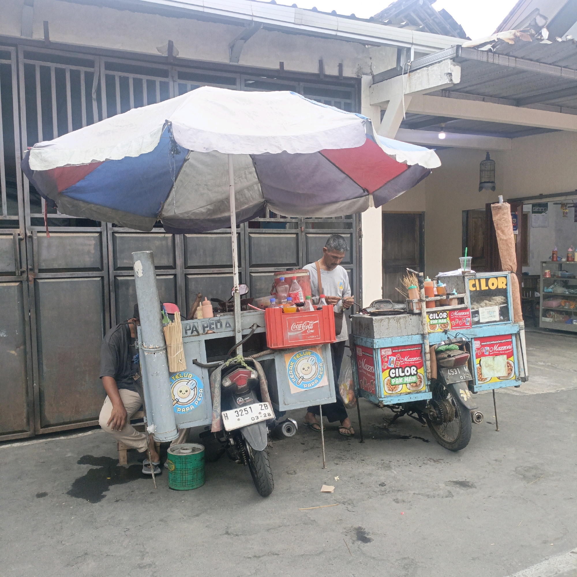 This is a daytime, eye-level, medium-shot photograph of a street food cart selling "Cilor" (a type of Indonesian fried snack) in front of a building with a metal gate in Salatiga, Indonesia. The cart is a modified motorcycle with a food stall structure attached. It is covered by a large, multi-colored umbrella, predominantly white, blue, and red.
Two men are present. One man, appearing to be the vendor, stands behind the cart, wearing a light gray t-shirt and a dark shoulder bag, and is in the process of preparing food with chopsticks. The second man, likely an assistant or customer, is seated on a wooden stool to the left of the cart, wearing a dark t-shirt and light brown pants. He appears to be resting.
The cart itself is adorned with colorful banners and logos, including "CILOR" and "MAZZONI". Several bottles of drinks, including Coca-Cola, and condiments are visible on the cart. A motorcycle is integrated into the cart's structure, with its license plate clearly visible as "H 3251 ZB 03-28". A green bin is placed on the ground near the seated man.
The background shows a building with a corrugated metal roof and a large, dark metal gate. Part of a bird cage is visible hanging from an overhang. The ground is paved with asphalt, with some damp patches and debris. The overall scene depicts a typical street-side food vending operation.