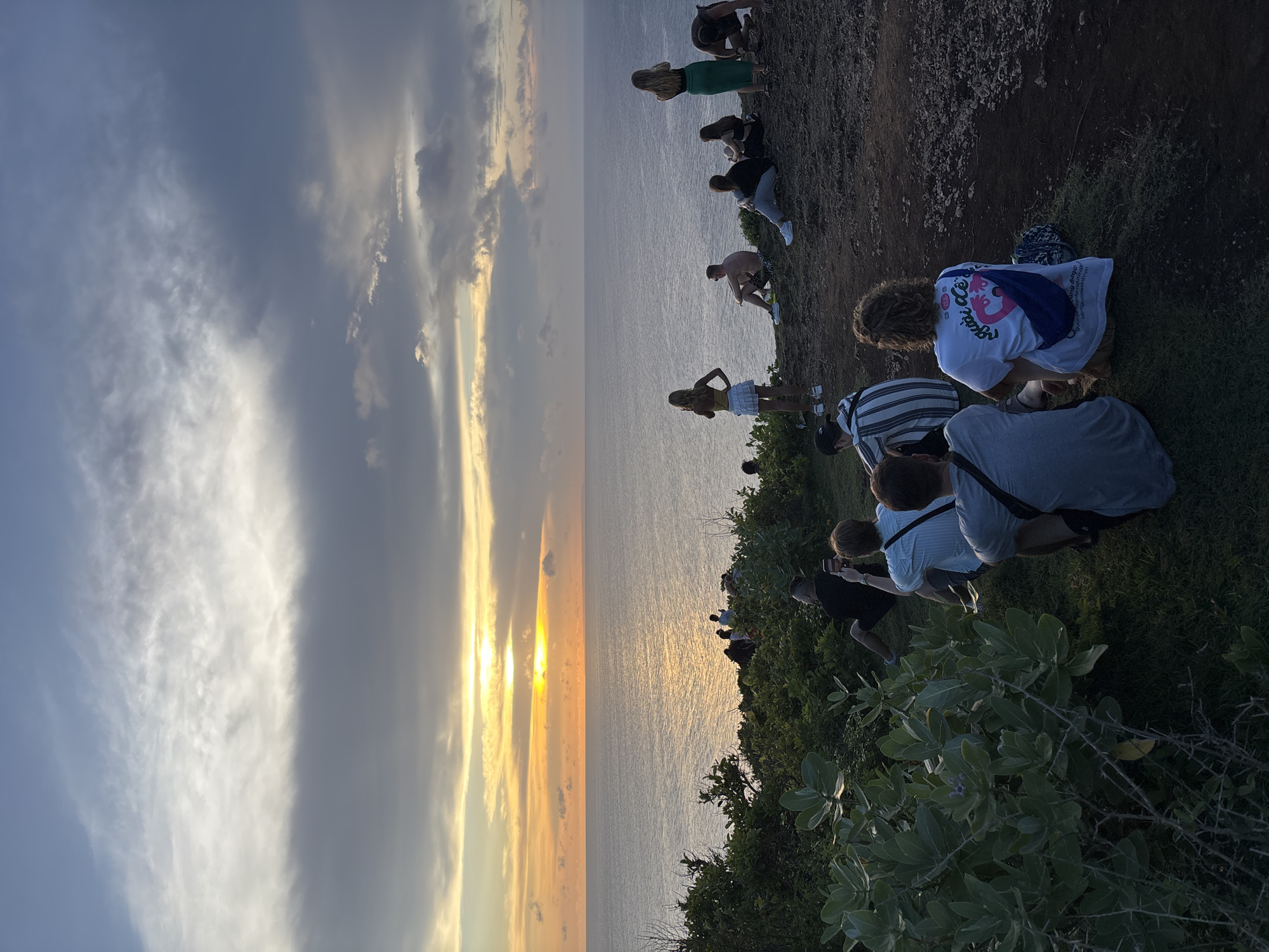 The image shows a group of people at Uluwatu, Indonesia, watching a sunset over the ocean. The sky is cloudy, with the sun visible through a gap in the clouds. The ocean is calm and reflects the colors of the sunset. The people are standing on a cliff, looking out at the view. Some people are taking pictures. The vegetation is green and lush near the cliff.