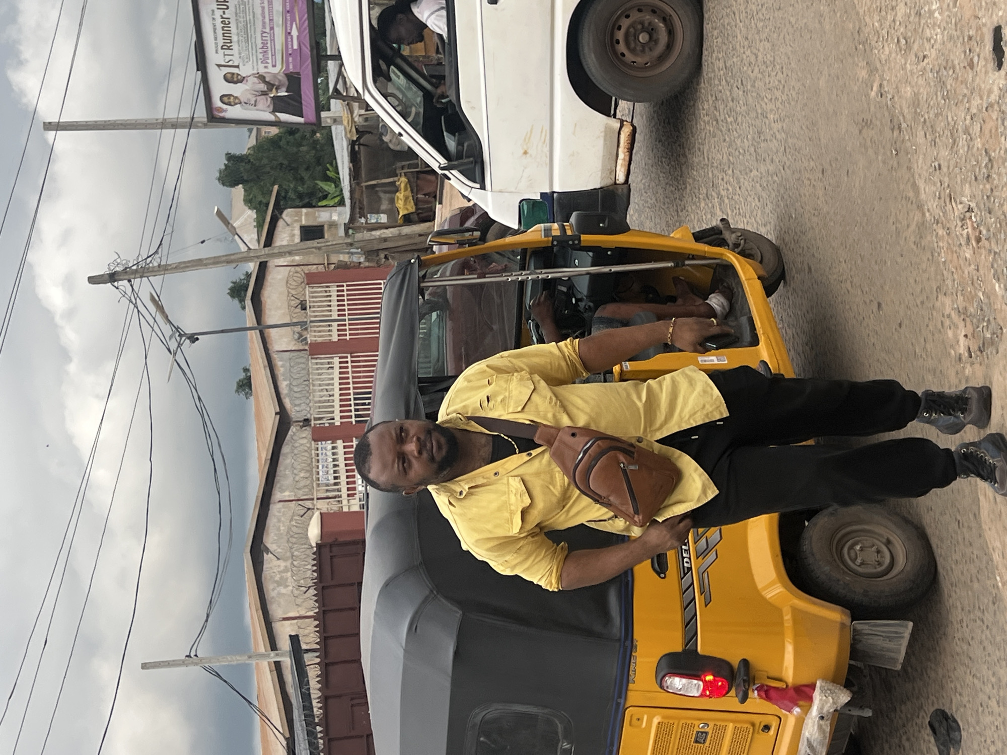 A man stands next to a yellow auto-rickshaw, wearing a yellow shirt, black pants, and black boots. He has a brown fanny pack slung across his chest. In the background, a white truck is parked, and buildings line a street. A billboard for "Pinkberry International School" is visible on the left side of the frame. The sky is overcast, suggesting it might be a cloudy day. The scene appears to be in a commercial or residential area. The man's expression is neutral, and he is looking towards the camera. The auto-rickshaw has the brand name "DELUXE" visible on its side. The setting suggests a typical day in Nkpor, Nigeria.