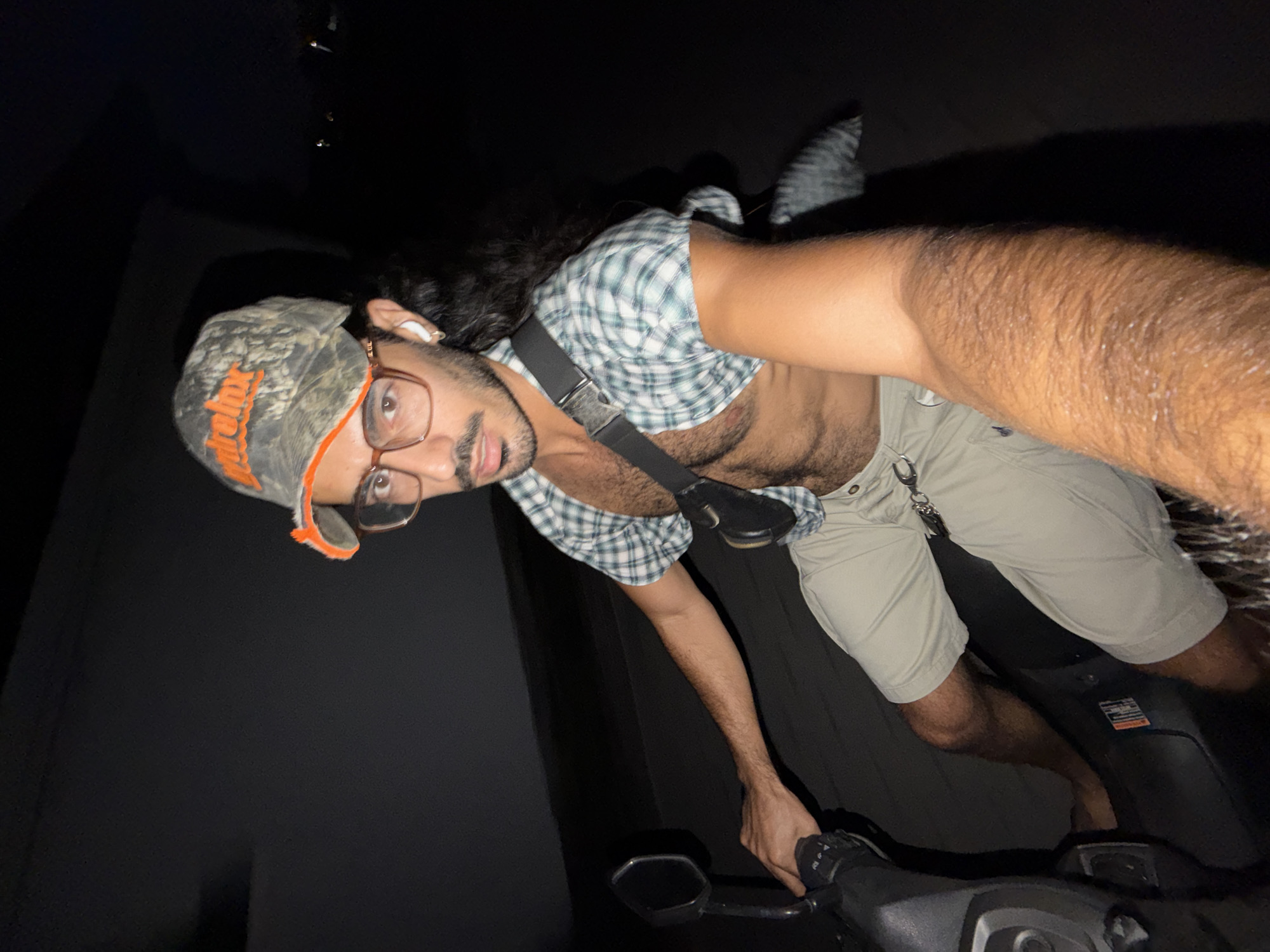 A person (male) takes a diagonal selfie while seated on a black scooter at night. He looks directly at the camera with a neutral expression. He wears a camouflage baseball cap with orange text, possibly "Tupelo," and light-framed glasses. A white wireless earbud is in his right ear. He has dark, curly hair and a beard.
He is dressed in a light-colored, short-sleeved plaid shirt, partially unbuttoned to reveal his hairy chest, and beige shorts. A black strap, likely for a bag or camera, crosses his chest, and keys hang from a loop on his shorts. His right arm is extended towards the camera, holding the device for the selfie, while his left hand rests on the handlebars of the scooter.
The background is completely dark, suggesting the photo was taken outdoors at night, with the subject brightly illuminated, likely by a flash. The handlebars and part of the scooter's body are visible in the foreground. The scene is set in North Kuta, Indonesia.