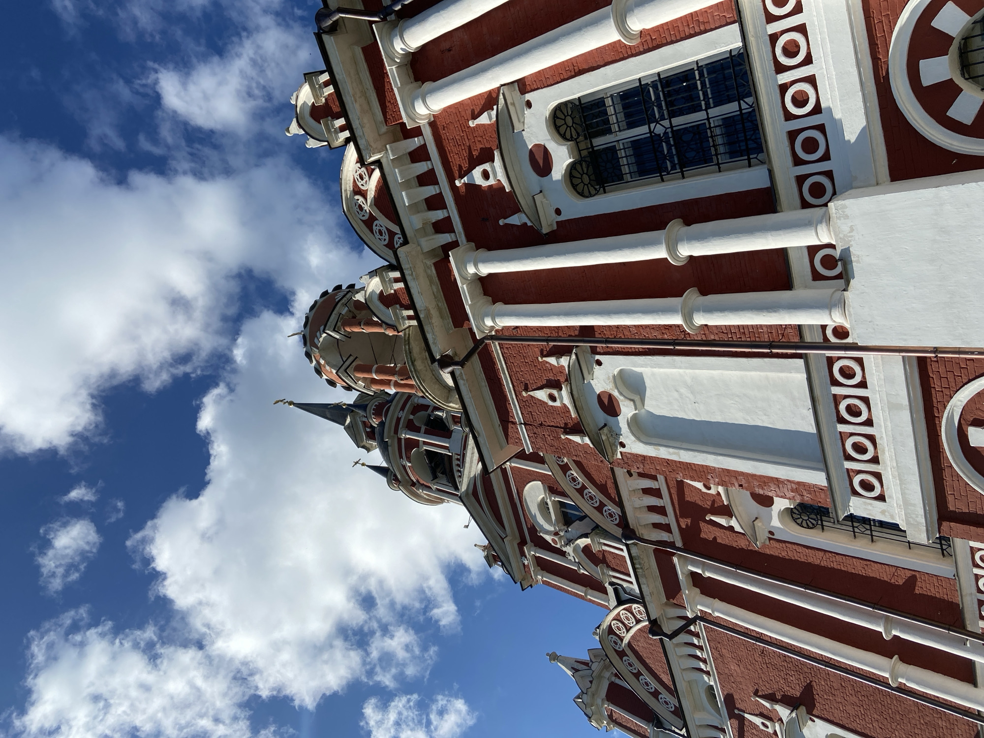 This is a low-angle, eye-level, exterior shot of the Church of St. Nicholas the Wonderworker in Mozhaisk, Russia. The church is constructed of red brick with white architectural details. It features multiple onion domes topped with crosses. The sky above is a vibrant blue with fluffy white clouds. The image focuses on the facade and upper structures of the church. There are no people visible in the frame.
