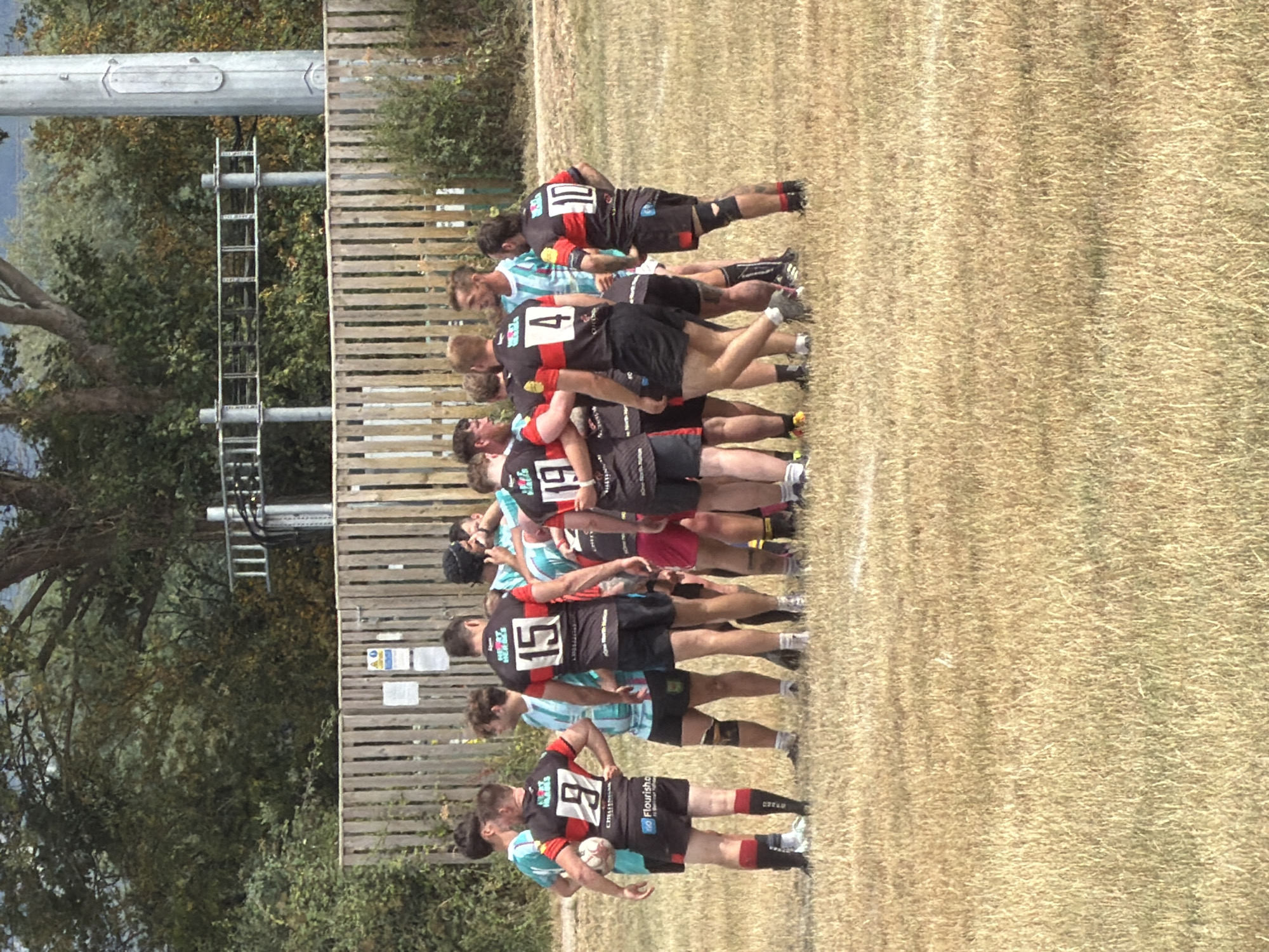 A group of rugby players, likely two teams, stand in a huddle on a dry, grassy field. The players are wearing jerseys with numbers and various colors, including black with red accents and light blue. Some players are holding a rugby ball. In the background, there is a wooden fence, trees, and some industrial-looking structures. The scene suggests a sports event taking place outdoors. The image does not provide enough context to determine the specific location within Tewkesbury, United Kingdom.