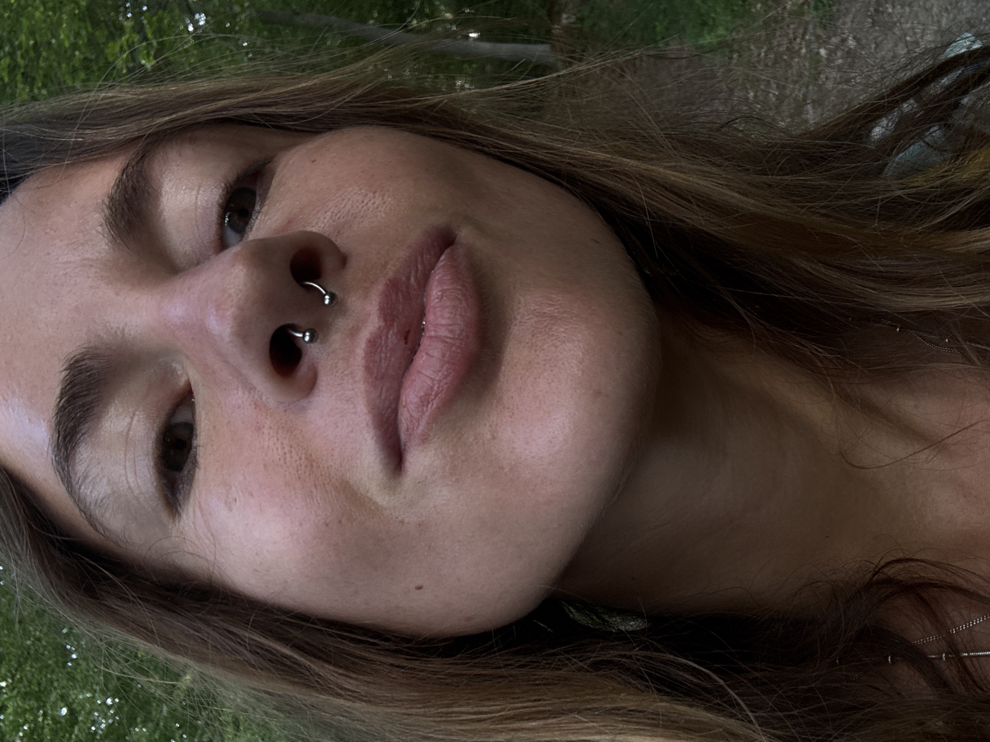 A close-up, low-angle, outdoor, selfie-style photograph shows a woman's face and upper chest against a blurred background of trees. The woman has long, light brown hair framing her face, and she is wearing a septum piercing. Her skin is visible with some pores and a mole on her cheek. Her mouth is slightly open, and her eyes are looking towards the camera. The lighting suggests it is daytime, possibly late afternoon or early evening, with some shadows indicating the direction of the sun. The environment appears natural, with greenery and tree trunks visible in the background.