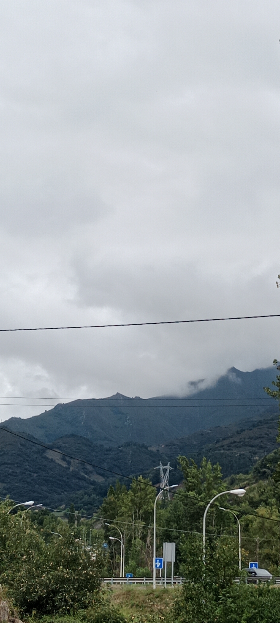 The image captures a view of a mountainous landscape under an overcast sky. In the foreground, dense green foliage is visible, with some trees and bushes. A road runs horizontally across the middle ground, bordered by a guardrail. Several streetlights are positioned along the road. Further in the background, the dark, tree-covered slopes of mountains rise, with some peaks shrouded in clouds. A tall electricity pylon is also visible on a distant mountainside. The scene is devoid of any people. The composition suggests a rural or semi-rural setting, possibly on the outskirts of a town like Lena, Spain, given the presence of infrastructure alongside natural scenery.