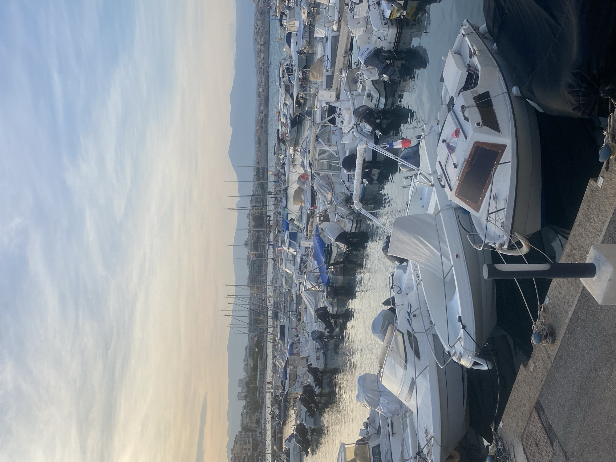 The image depicts a marina in Antibes, France, filled with numerous docked boats. The sky above is a soft gradient of pale blue and yellow, suggesting either sunrise or sunset, with distant mountains forming a hazy backdrop. The water is calm and reflects the boats and the sky. Most of the boats are small to medium-sized, many with their engines visible. Some are covered with tarps, while others are exposed. In the background, a town with buildings and more masts of sailboats can be seen. The foreground shows a concrete pier with mooring lines and cleats. There are no visible people in the image. The overall scene is one of a quiet harbor at either dawn or dusk.