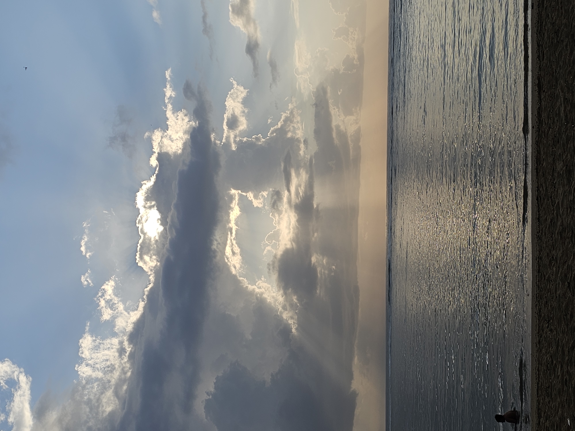 This is a vertical, medium-shot photograph taken at sunset on a beach in Denpasar, Indonesia. The sky is filled with dramatic clouds, with shafts of sunlight breaking through from behind them, illuminating the ocean. The ocean is a deep blue-gray, with gentle waves lapping at the shore. The sand in the foreground is dark and wet. In the shallow water near the shore, a person is visible from the shoulders up, facing away from the camera and towards the ocean. The overall mood is serene and tranquil.