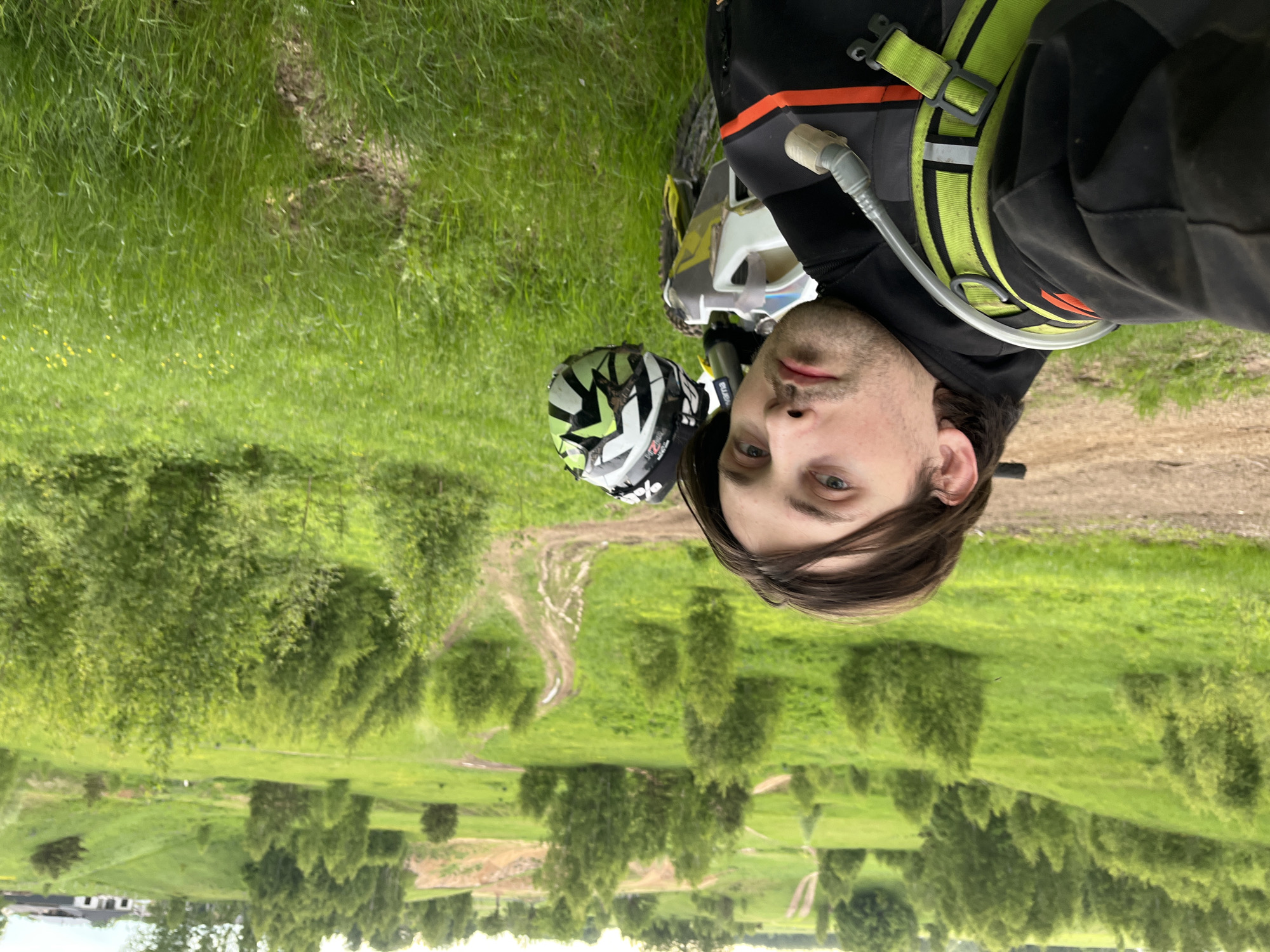 A man with long dark brown hair, wearing a black hooded jacket, a neon green and black hydration pack, and standing next to a motorcycle with a helmet, is photographed outdoors in a grassy field in Новочувашский, Russia. The photo appears to be taken from a slightly upward angle. A dirt trail is visible in the background, winding through the landscape dotted with small trees and vegetation. In the distance, buildings can be seen.