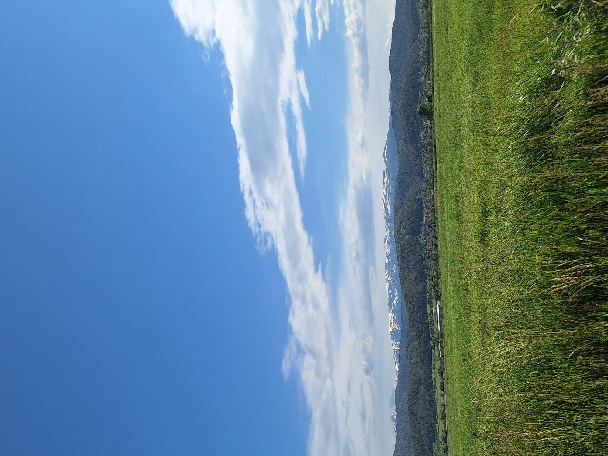 The image depicts a landscape with a field of green grass in the foreground, likely in a rural setting, not specifically identifiable as Alino, Bulgaria without further context or identifiers. A mountain range with some visible snow on the peaks runs along the middle ground. The sky occupies a significant portion of the image, displaying a clear blue color with long, white clouds stretched across it. There are no visible people, actions, or interactions. The environment appears to be a sunny, outdoor location. The dominant subjects are natural elements: the grass, mountains, and sky. An object or structure, possibly a building, can be seen in the middle ground near the mountains.