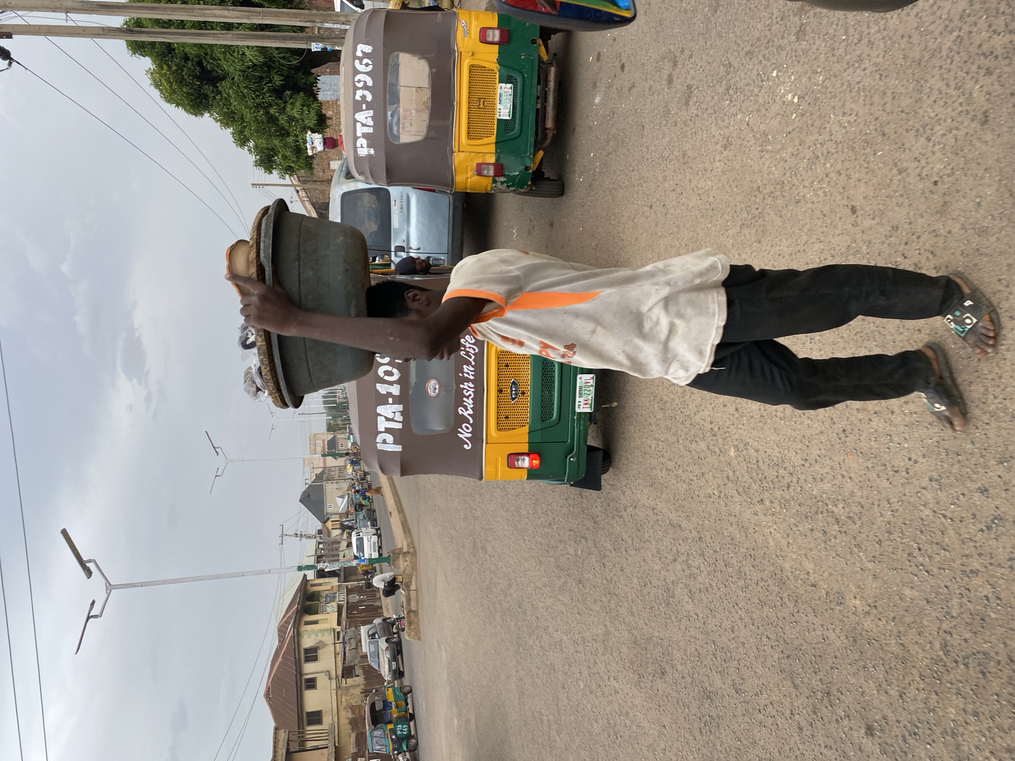 The image shows a man in Bukuru, Nigeria, carrying a large basin on his head. He is wearing a white shirt with an orange stripe and black trousers. The man is walking on a street lined with auto rickshaws known as "Keke Napep," each having different registration plates. One has the inscription "No Rush in Life" on it. There are buildings on the side of the street, as well as power lines overhead. The sky is overcast.