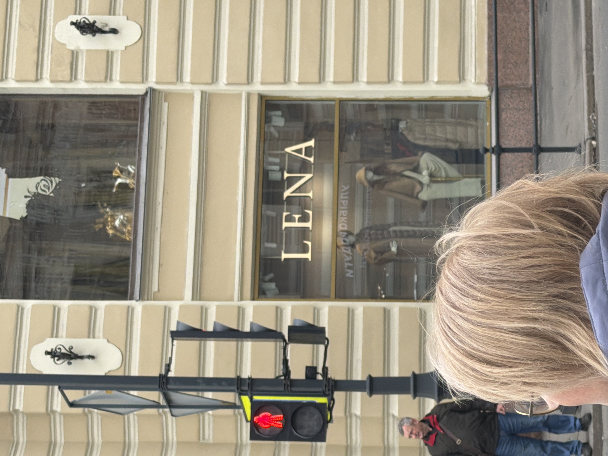 This image captures a vibrant street scene, likely in a commercial area of Saint Petersburg, Russia, featuring a building with a storefront, a pedestrian, and a traffic light. The photograph appears to be rotated 90 degrees counter-clockwise, with the building facade extending horizontally and the street vertically.
In the foreground, the back of a person's head and shoulder are visible. This individual has long, light blonde hair and is wearing a blue jacket. Their head is positioned in the lower right portion of the rotated image.
The prominent feature is a building with a light-colored, vertically striped facade. On its ground floor are two large display windows, likely belonging to a shop. Inside the right-hand window, mannequins are visible, dressed in various coats and attire, reflecting the nature of a clothing store. The shop's name, "LENA," is prominently displayed in large, gold, serif letters on this window. Below "LENA," smaller Cyrillic text is visible, partially obscured by reflections, appearing to read "АЛЛЕГРО. ОДЕЖДА" (Allegro. Clothes), further confirming the type of establishment. Decorative, ornate wall sconces are mounted on the building's facade, one above each window.
In the middle-ground, a black traffic light pole extends horizontally across the frame (when oriented correctly). Suspended from it is a pedestrian signal showing a red "don't walk" figure.
Further in the background, a man with gray hair is visible, wearing a dark jacket, a red scarf, and blue jeans. He appears to be walking on the sidewalk. The scene takes place during daytime, indicated by the clear and bright lighting. No specific weather conditions are discernible, suggesting a typical clear day. The architectural style, combined with the Cyrillic text, aligns with the provided location context of Saint Petersburg.
