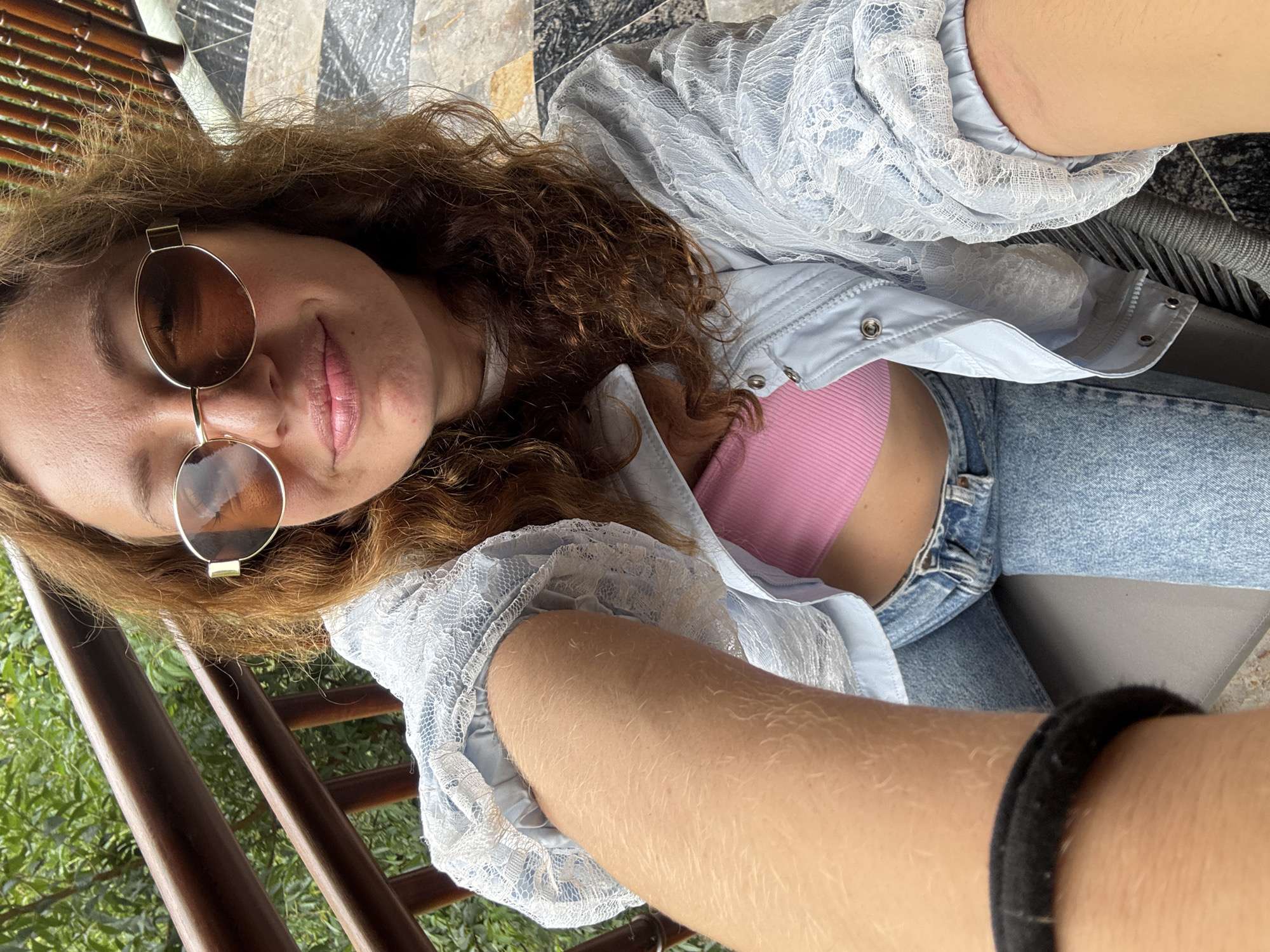 A woman is captured in a close-up, high-angle selfie. She has light brown, curly hair and wears brown-tinted sunglasses with gold-toned frames. Her lips are slightly parted in a faint smile. She is dressed in a light blue jacket with white lace detailing on the sleeves and shoulders, worn over a pink ribbed crop top, and blue denim jeans. Her left arm, positioned in the foreground and extending towards the camera, shows fair skin with visible light hair, and a black fabric band is present on her wrist. The background features a wooden railing on the left, beyond which is lush green foliage. On the right, a structure of vertical wooden slats is visible, adjacent to a patterned tiled floor in shades of grey and black. The scene depicts an outdoor setting, located in Púng Luông, Vietnam.